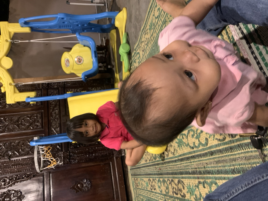 The image displays an indoor or semi-enclosed scene in the city of Mojorejo, Indonesia, featuring two children and partial view of an adult, surrounded by play equipment and furnishings.
In the foreground, a baby or toddler, wearing a light pink ruffled top and blue jeans, is positioned on a green and light brown patterned mat. The child's upper body, profile of the face, and part of the head are visible, facing towards the right with eyes looking slightly upwards. Small gold stud earrings are present. To the far left, a portion of an adult's leg, clad in blue denim, is visible on the mat. A dark object, resembling a car key or remote, lies on the mat near the adult's leg.
In the mid-ground, a young girl with dark, straight bobbed hair is seated cross-legged on a yellow plastic component of a playset. She wears a bright pink collared short-sleeved shirt and gazes directly at the camera.
Behind the children, a multi-component plastic playset, predominantly yellow and blue, occupies the space. It includes a yellow slide with a blue frame on the left, to which a small white-netted basketball hoop with an orange rim is attached. To the right, a blue-framed swing set holds a yellow bucket swing seat, which features a discernible "Burger King" logo. A green plastic toy lies on the mat near the base of the swing set. The background consists of dark brown, intricately carved wooden furniture, possibly a cabinet or screen. The floor is entirely covered by the patterned mat with striped and floral designs.