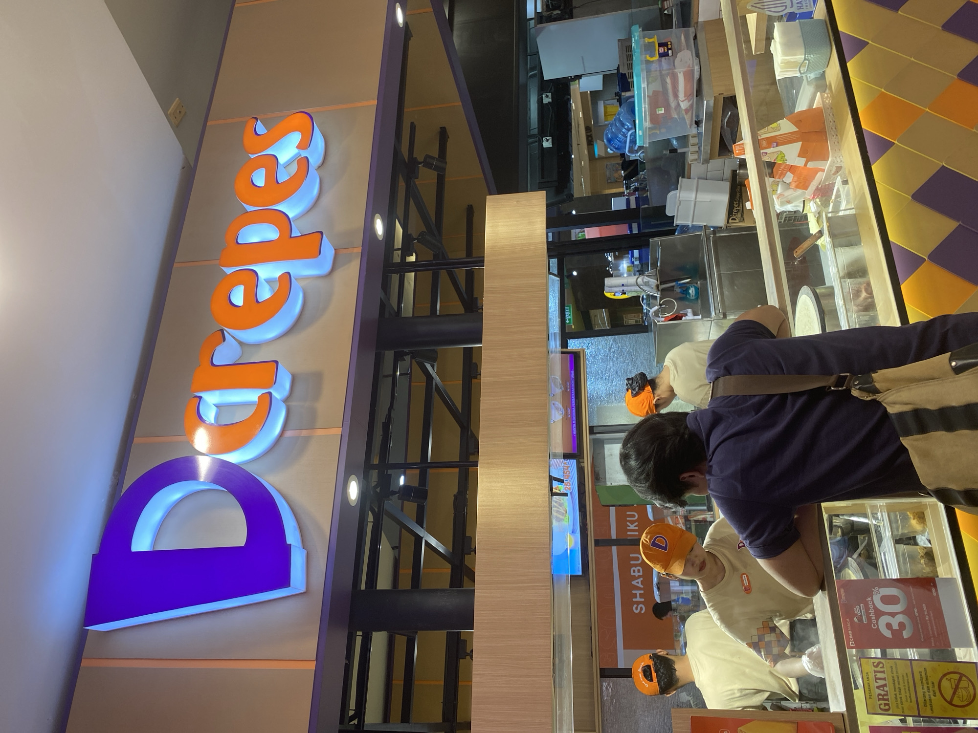 In West Jakarta, Indonesia, at a Darepes crepe shop, a man wearing a navy blue polo shirt and a striped bag over his shoulder is looking at a crepe station. Behind the counter, three employees, two wearing orange baseball caps and one with an orange cap with a "D" logo, are preparing food. One employee, a woman in a beige shirt and an orange cap, is interacting with the customer. The shop features a prominent purple and orange "Darepes" logo illuminated with blue light. Displays promoting "Gratis" (free) and "Cashback 30%" are visible, indicating a promotional offer. The environment appears to be a food court or mall setting.