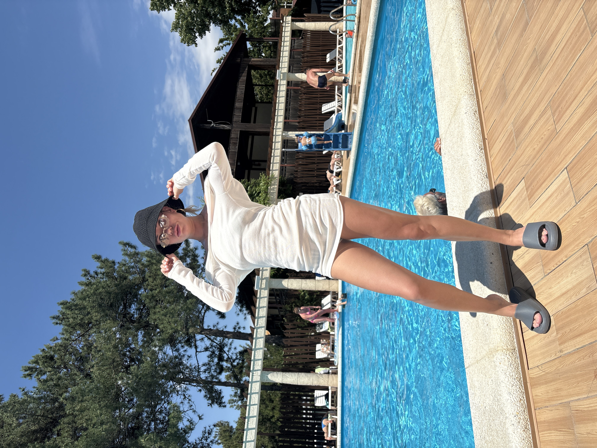 The image depicts a sunny day at an outdoor swimming pool, possibly in Anapa, Russia, given the context. In the foreground, a woman stands on a wooden deck, wearing a white off-the-shoulder dress, sunglasses, and a black bucket hat. She is holding the brim of her hat with both hands. She is also wearing grey slide sandals. The pool behind her is filled with bright blue water. Several people are visible around the pool, some lounging on chairs, others in the water, and a child is on a blue slide. A wooden structure with an overhang is visible in the background, as are trees and a clear blue sky. The overall scene suggests a recreational setting.