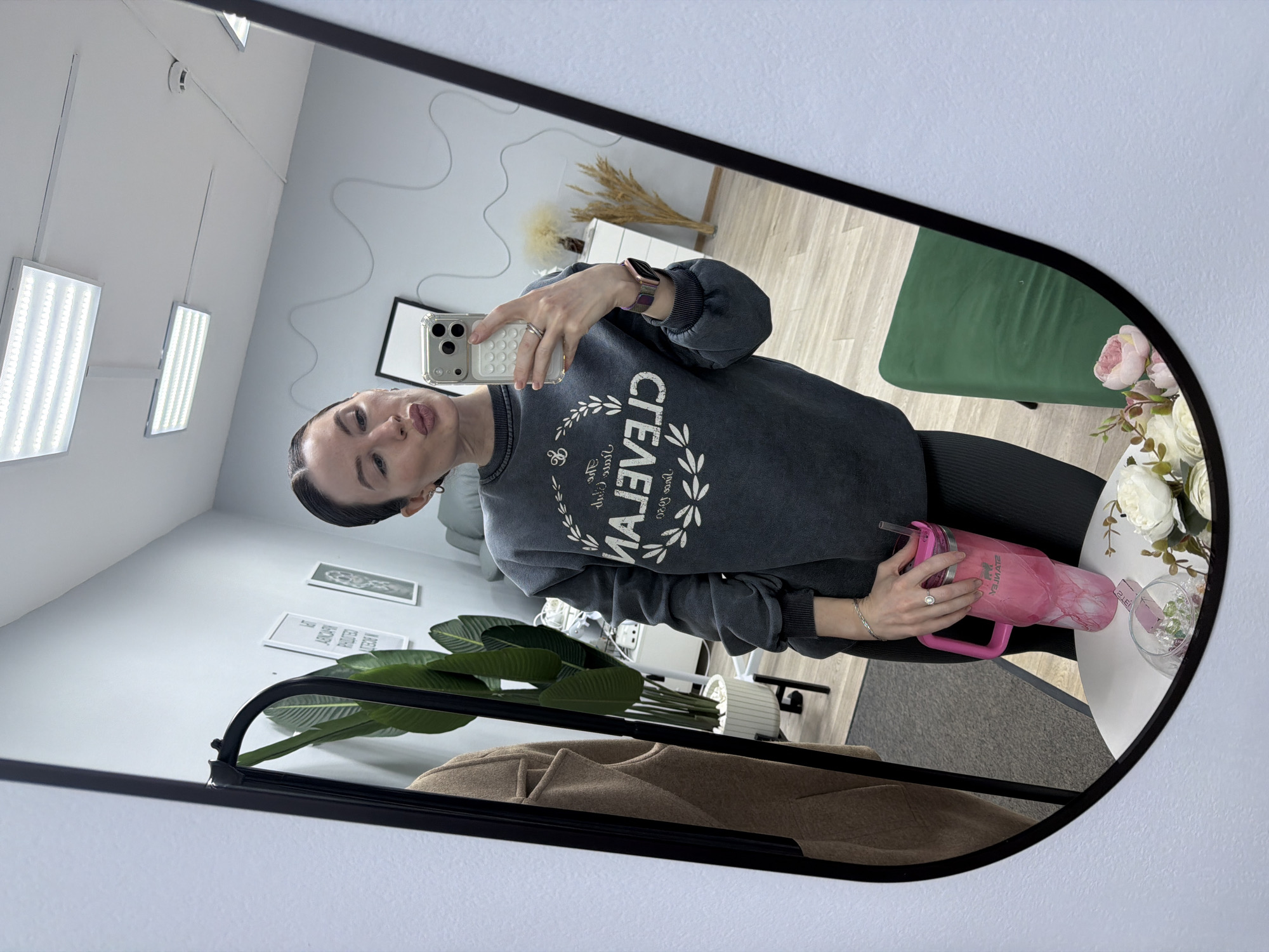 A woman is captured in a mirror selfie, standing indoors in what appears to be a modern commercial or office space. She is holding a smartphone with a clear, textured case in her right hand to take the picture, and a pink, marble-patterned "STANLEY" brand tumbler with a handle in her left hand.
The woman wears a dark grey, washed-out sweatshirt with "CLEVELAND" printed across the chest in white, accompanied by a laurel wreath design and smaller, mostly illegible text below it. She is also wearing black leggings, a watch on her left wrist, and rings on her right hand. Her expression is neutral, with slightly pursed lips.
The reflection in the large, black-framed mirror reveals a bright and clean room with light-colored walls and a light wood-look floor. The ceiling is illuminated by two rectangular LED light panels. Decorative elements in the room include a wavy black line on one wall, framed pictures (one with the word "PERM" visible), and a large artificial plant with broad green leaves. Also reflected are a green upholstered chair and a small round white table decorated with an arrangement of white and pink artificial flowers, primarily peonies, and a glass dish holding colorful decorative beads or stones. A beige coat or jacket is visible, hanging on a rack in the lower part of the reflection. The location context is Perm, Russia.