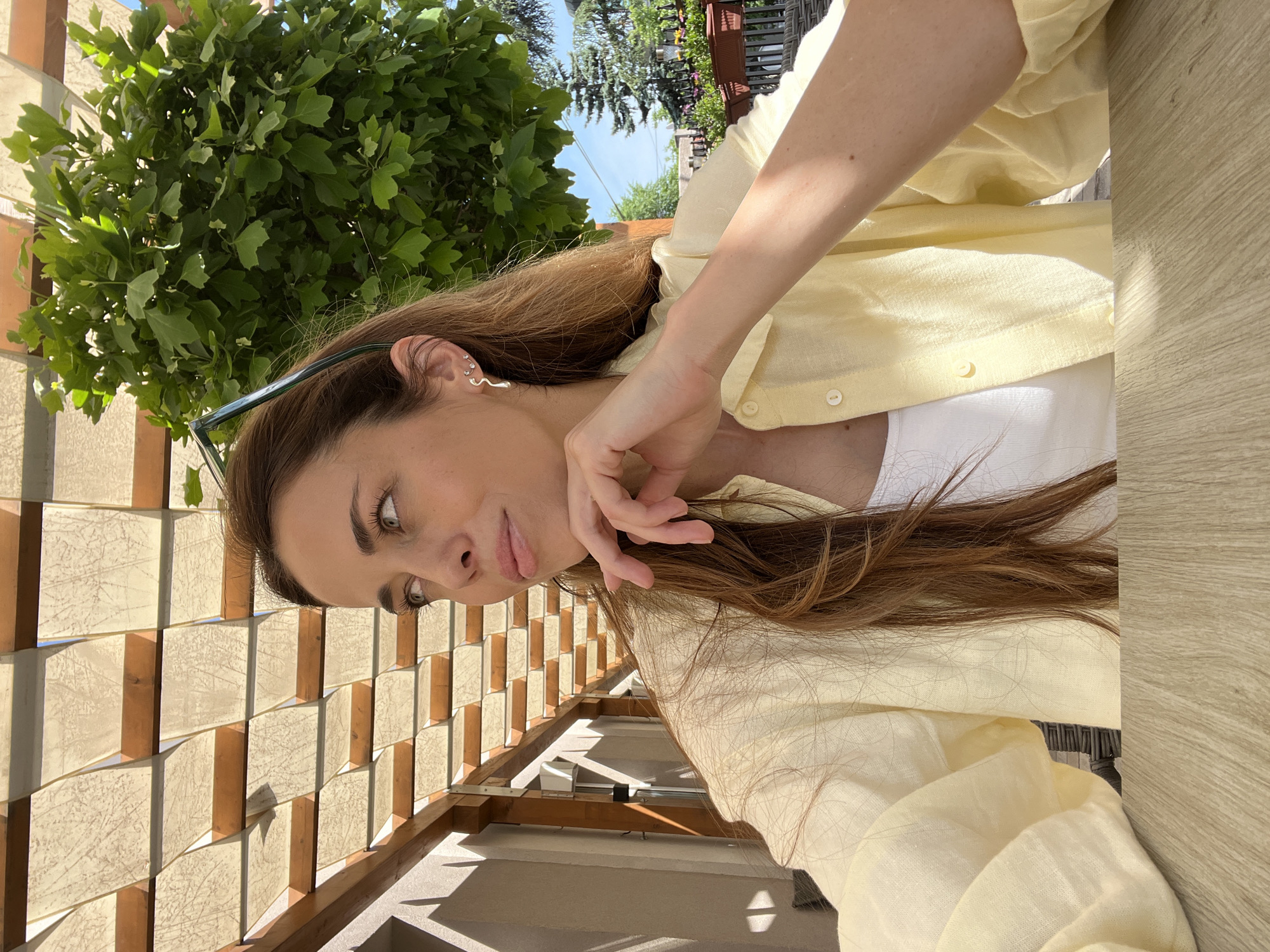 A woman is captured in an outdoor setting, possibly in the Vracar Urban Municipality, Serbia. She is positioned near a decorative wall composed of beige and brown rectangular blocks, suggesting a modern or contemporary architectural style. The woman's attire consists of a light yellow button-up shirt over a white undershirt. She wears sunglasses, and a distinctive silver serpent earring is visible in her left ear. Her hair is brown and appears well-kept. The overall ambiance suggests a sunny day, with natural light casting shadows and illuminating the scene. Green foliage is visible in the background, hinting at nearby trees or landscaping.
