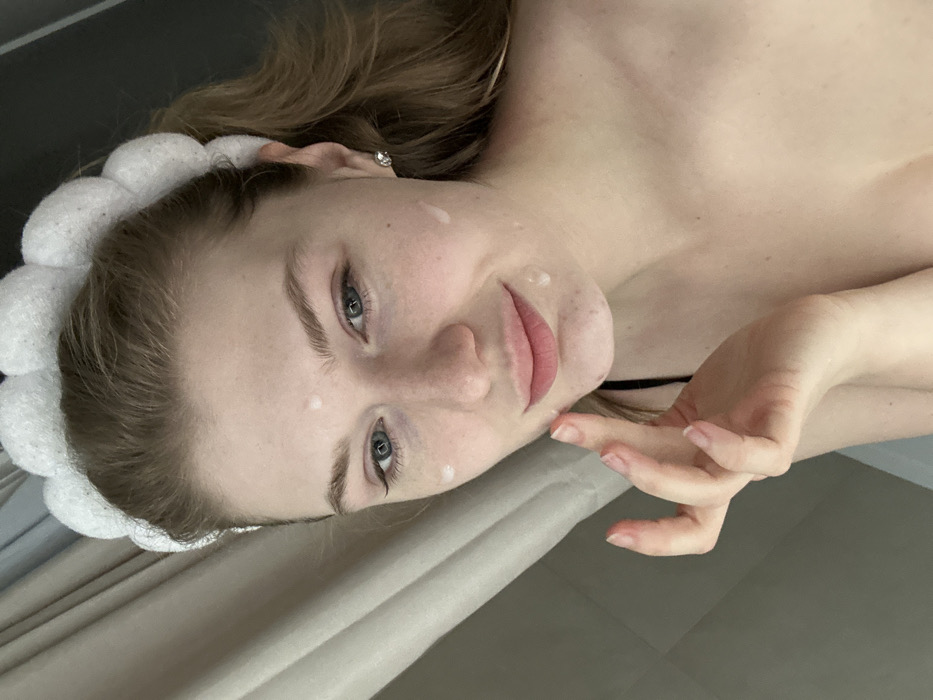 A pale-skinned woman lies down, facing the camera. She wears a white, plush headband. Her eyes are blue, and her lips are pink. Several white, opaque droplets are visible on her forehead and cheek. Her right hand is raised near her face. A thin, black strap is visible at her neck. The background includes a textured cream-colored surface. The image is rotated to the left.