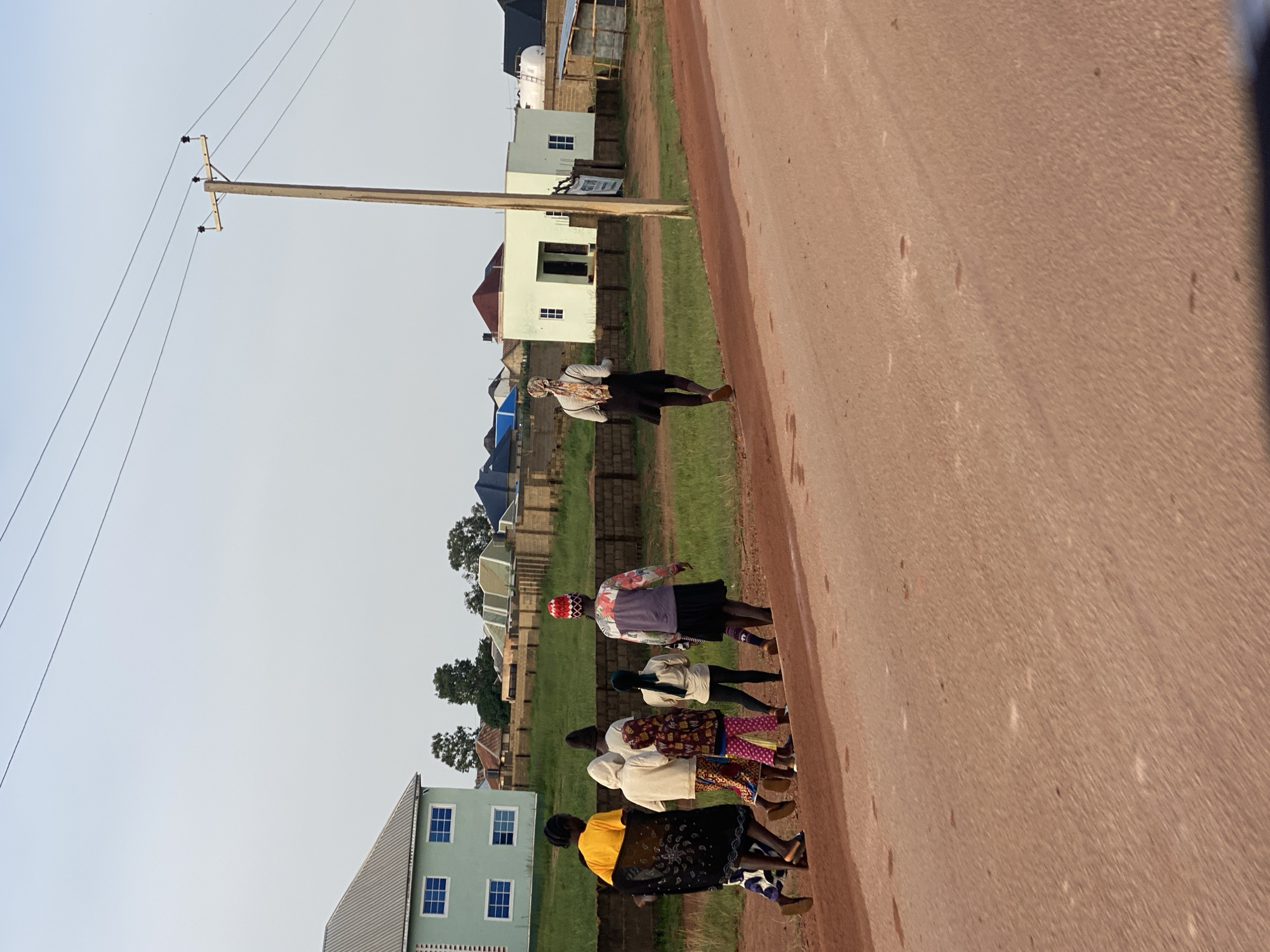 In the city of Kwang, Nigeria, a group of seven individuals, appearing to be women and children, walk along a dirt road. The group is moving from left to right in the frame. The person at the front of the group on the left wears a bright yellow top and a patterned black wrap. The individuals behind her are dressed in various colorful clothing, including a white hoodie, patterned pants, and a vibrant red and white head covering. The person furthest to the right in the group wears a patterned jacket over a darker skirt.
In the background, several buildings are visible. A light green, two-story building with blue-trimmed windows is on the far left. Other buildings, some with dark tiled roofs and some with lighter colored walls, are scattered throughout the scene. A wooden utility pole with power lines extends from the right side of the frame towards the center. The ground is predominantly a reddish-brown dirt, with patches of green grass appearing between the road and the buildings. The sky is a pale blue with a hazy appearance. The lighting suggests it is either early morning or late afternoon.