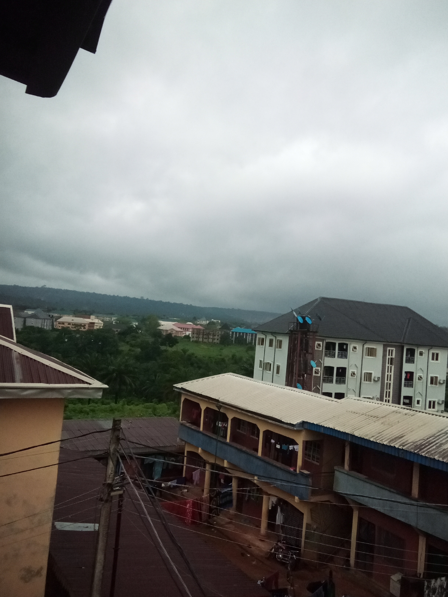 The image captures an overcast day in Amaokpala, Nigeria, showcasing a mix of residential and commercial buildings set against a backdrop of lush greenery and distant hills. In the foreground, a light-colored building with a corrugated metal roof is partially visible on the left, with electrical wires crisscrossing in front of it. Adjacent to this, a two-story structure with yellow pillars and a light-colored metal roof features a blue trim along its upper level, where laundry is hung out to dry. Below this, another building with a darker, corrugated metal roof is visible, with a motorcycle parked in front of it.
Further into the scene, a large, multi-story building with a dark gray, pitched roof dominates the mid-ground. This building has a light-colored facade with numerous windows and balconies. To the left of this building, more structures of varying heights and colors, including one with a pink roof, are interspersed with dense green foliage. The background is characterized by a chain of hills covered in vegetation, receding into a hazy, gray sky. The overall impression is one of a densely populated area with a tropical climate.
