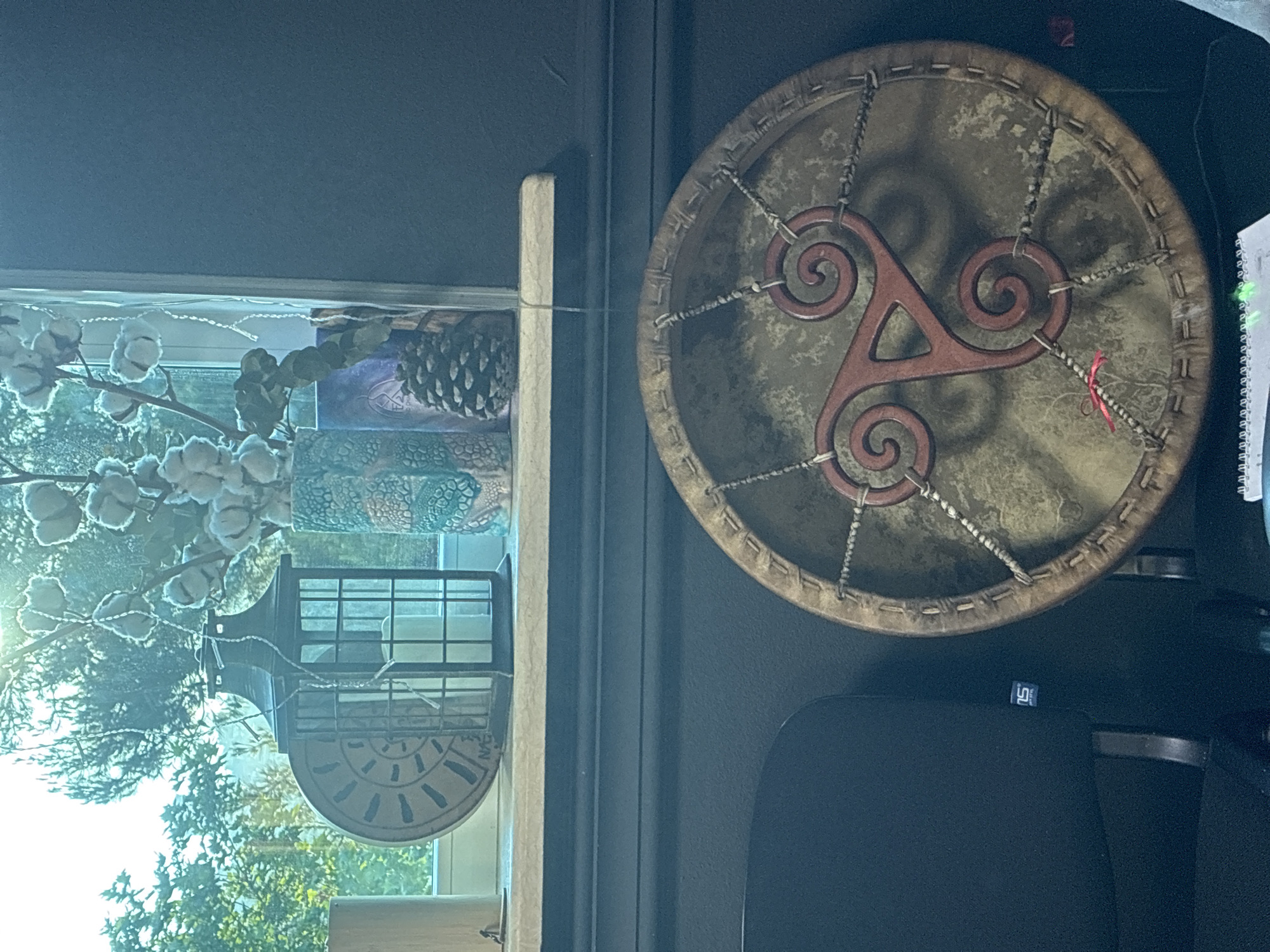 The image shows a decorative drum with a red triskelion symbol painted on its surface. It is placed on a dark surface, possibly a table or a cabinet. In the background, a window is visible with a shelf above it. On the shelf, there is a decorative lantern, a vase with cotton branches, and a pine cone. The window looks out onto a green, leafy area. There is no clear indication of the location within the city of Sochi, Russia.