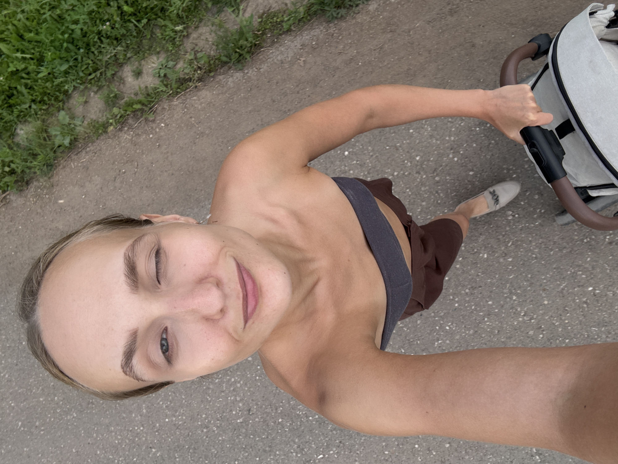 A woman is taking a selfie from a high angle, winking at the camera. She has fair skin, blonde hair pulled back, and is wearing a dark grey bandeau top and brown shorts. She is holding onto the handle of a stroller with her right hand. The stroller is light grey with brown accents. She is standing on a paved surface that appears to be a path or sidewalk, with a strip of green grass to her right. The scene suggests an outdoor setting in the village of Podshivalovo, Russia, possibly a park or residential area, given the presence of a stroller and greenery.