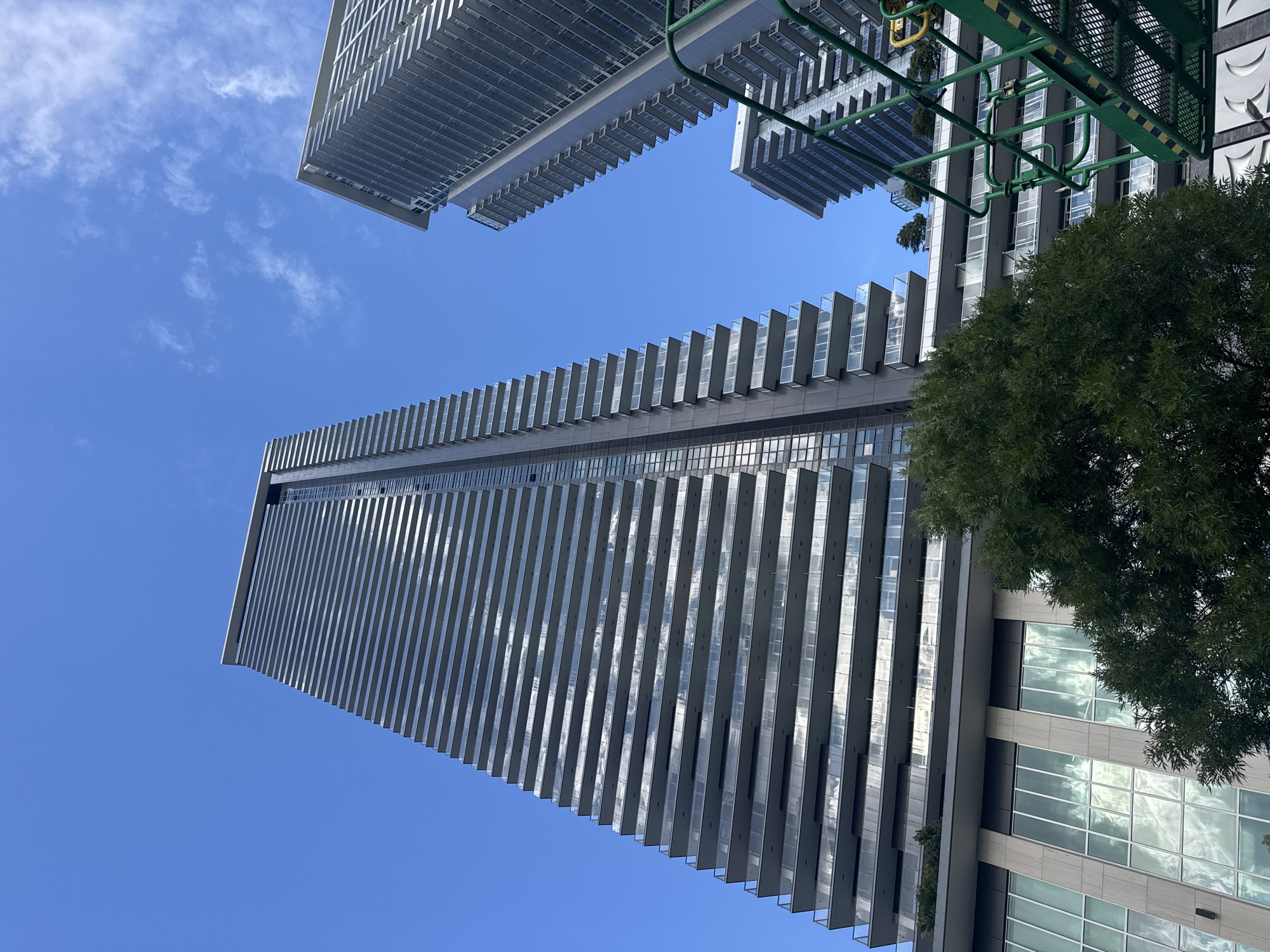 The image shows a low-angle view of modern high-rise apartment buildings against a clear blue sky with scattered clouds. The buildings feature repetitive horizontal balconies with glass railings, giving them a layered appearance. The closest building on the left is a very tall skyscraper, while another building of similar design is visible to its right, further in the background. A smaller, lighter-colored building with balconies is also present in the middle ground. In the foreground, a large green tree partially obscures the lower section of the closest building, which has a base with large, reflective windows. A green industrial lift or platform is visible in the lower right corner of the image, suggesting ongoing construction or maintenance. The scene appears to be an urban environment, likely Burnaby, Canada, given the architectural style common in the city. No people are visible in the frame.