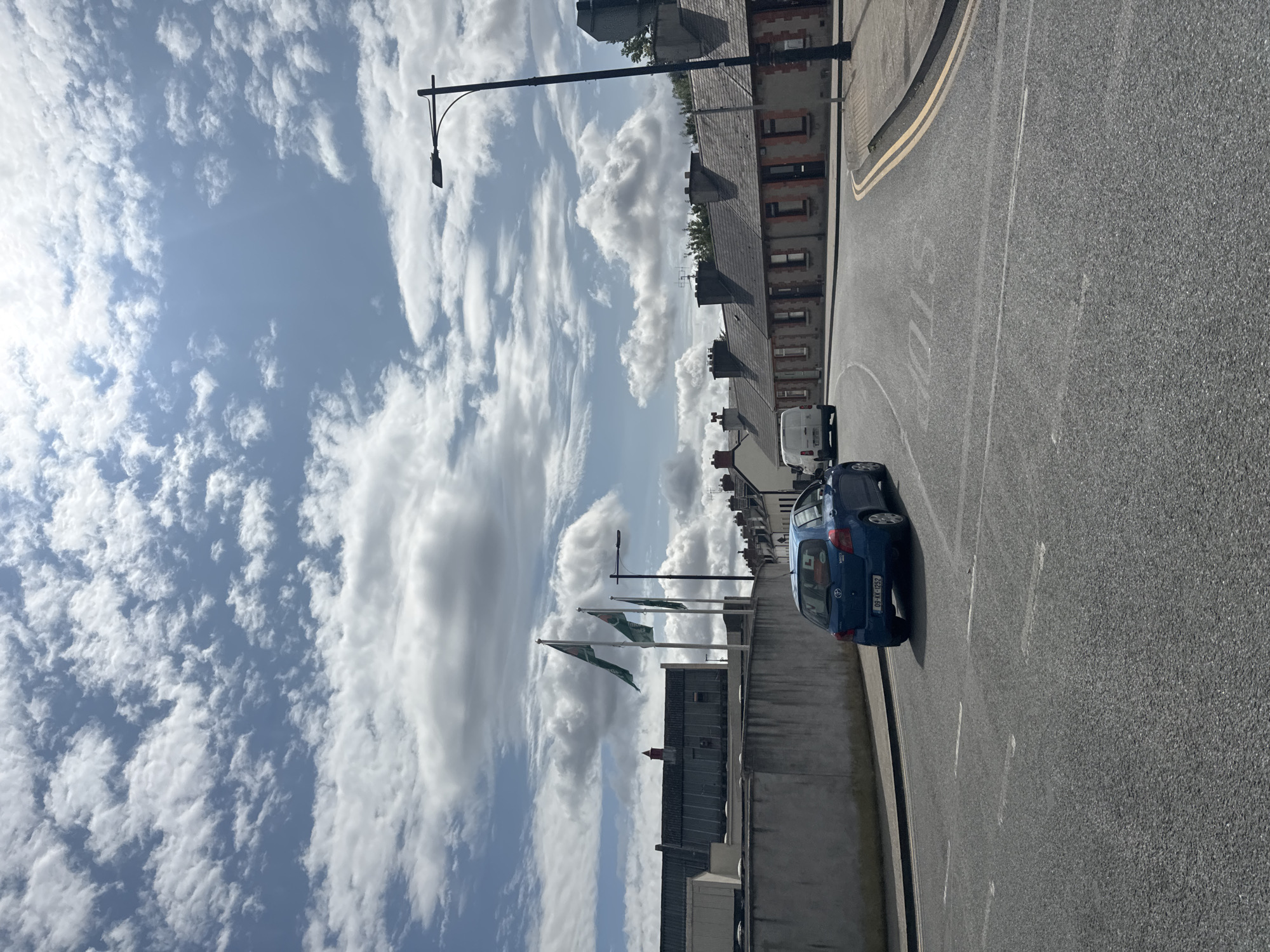The image depicts a street scene in Kilkenny, Ireland, under a partly cloudy sky. In the foreground, a blue Toyota Yaris is stopped or moving slowly on a paved road marked with white lines. The car displays an "L" plate, indicating a learner driver. Further down the road, a white van is visible. On the left side of the road, a grey concrete wall runs alongside a building with a dark roof. On the right side of the street, a row of terraced houses with grey tiled roofs and brick facades is present. Three green flags are flying from tall poles behind the concrete wall, with a lamppost and a street light positioned in front of the houses. The sky is filled with scattered white clouds against a light blue backdrop, with the sun partially obscured by clouds, casting light and shadows. The overall scene suggests a typical day in a residential or commercial area of Kilkenny.