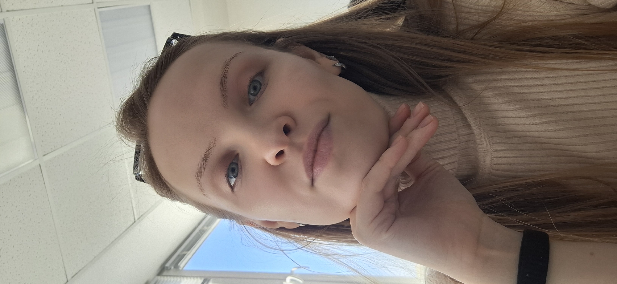A young woman with long brown hair and blue eyes is posing for a selfie indoors. She is wearing a light beige turtleneck sweater. Her left hand is resting on her chin. She is wearing a black wristband. Behind her is a white tiled ceiling and a window showing a blue sky. The location is Chaikovsky, Russia.