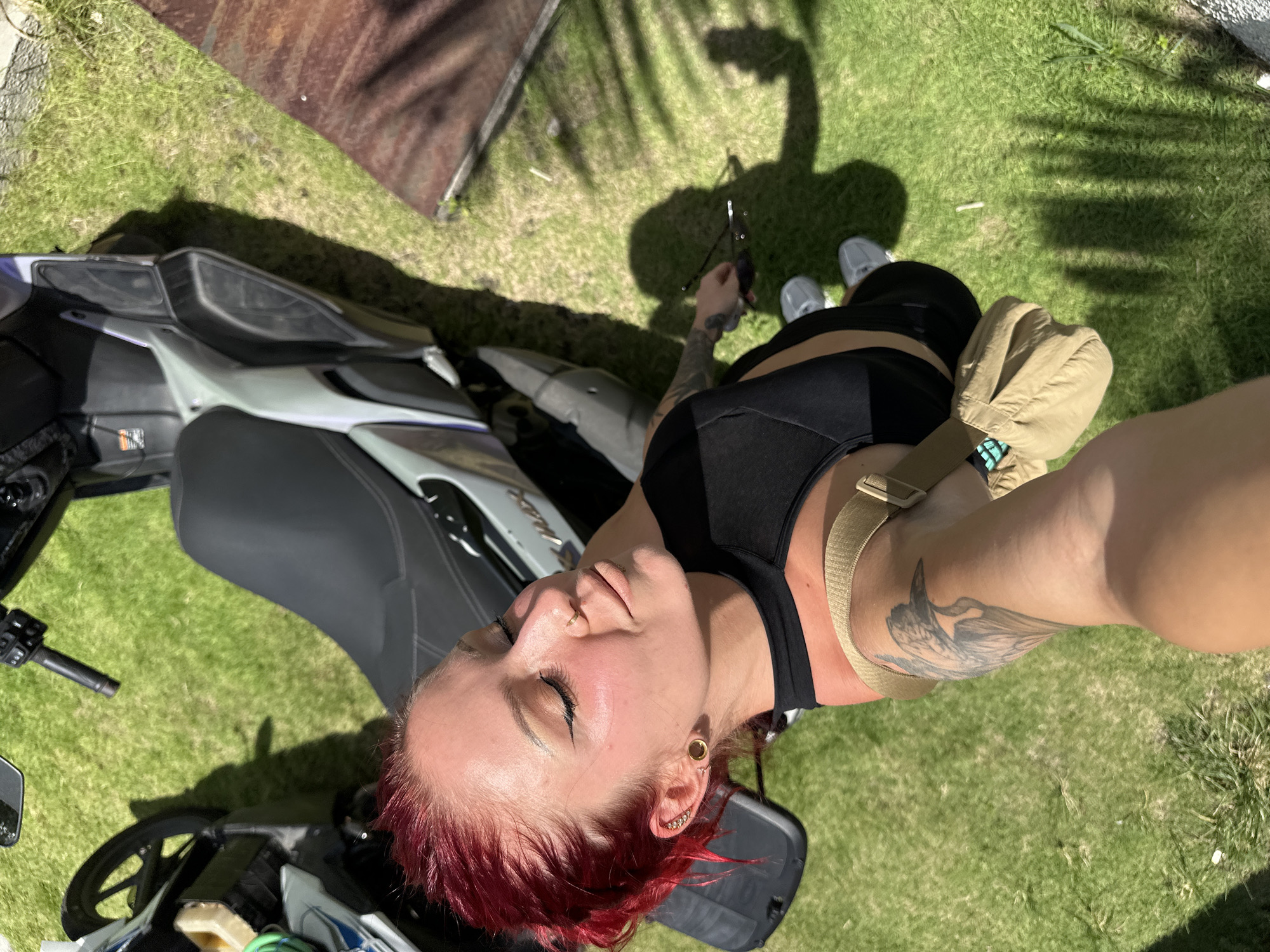 A medium shot of a red-haired person wearing a black top, black shorts, and gray sneakers standing on a green lawn. The person is adjacent to a parked motorcycle. The location is identified as Dalung, Indonesia.