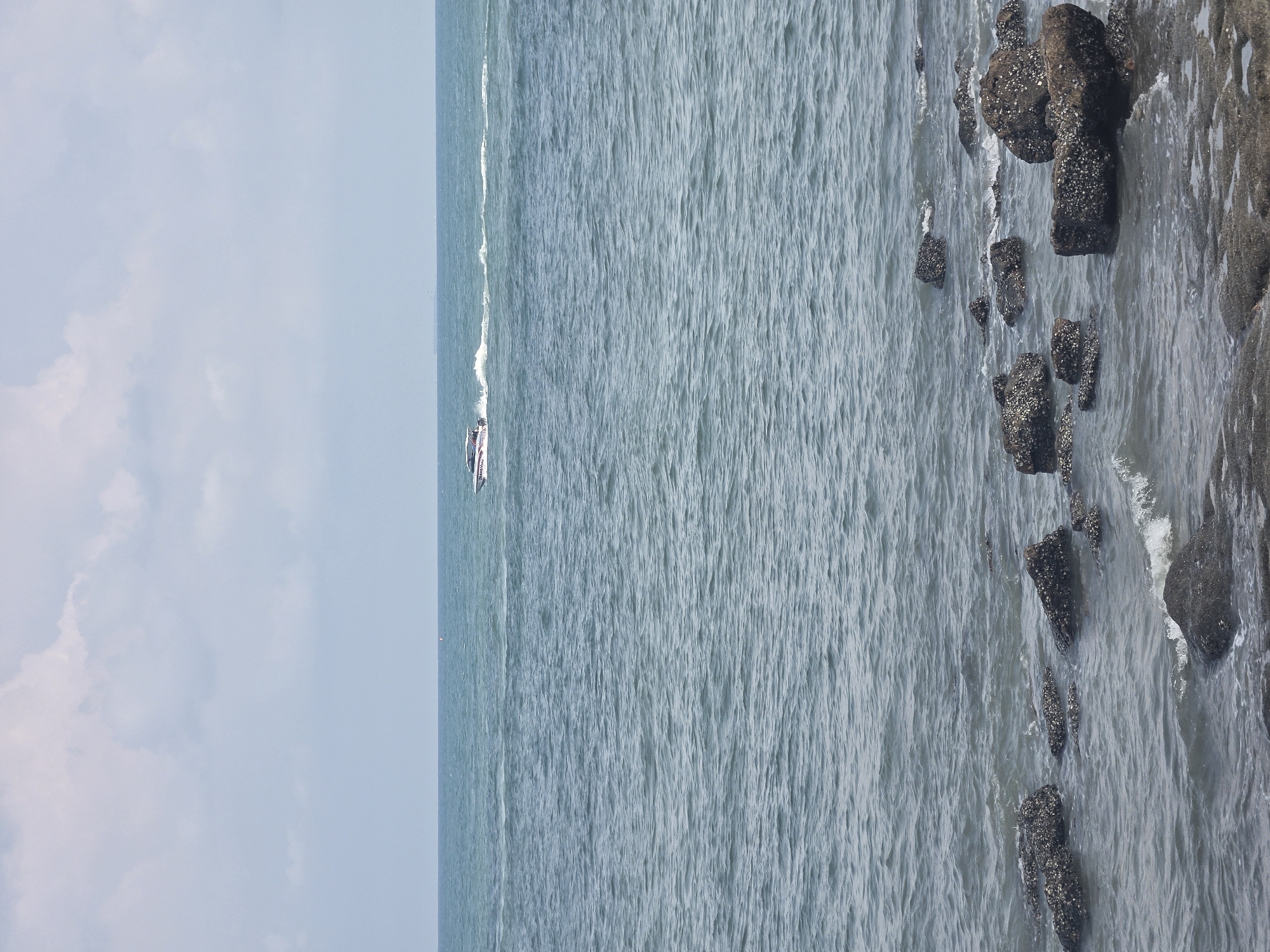 The image displays a coastal scene, likely in Pattaya City, Thailand, featuring the sea, a boat, a rocky shoreline, and the sky. The expansive blue-green ocean dominates the mid-ground, showing light ripples and texture from small waves. A white powerboat or speedboat, possibly a catamaran, is seen cruising from left to right across the water, leaving a clear white wake behind it. Red markings are visible on the boat's side, though any specific text is illegible due to distance.
To the right, a rugged shoreline is visible, characterized by numerous dark, irregular rock formations partially submerged in the water. These rocks are encrusted with barnacles or similar marine growth. Gentle waves lap against the rocks, creating subtle white foam. The sky above is light blue with scattered, soft white clouds, suggesting clear weather. The overall impression is a serene daytime view of a coastal area, likely a popular spot for boating and recreation. No people are discernible in the frame.