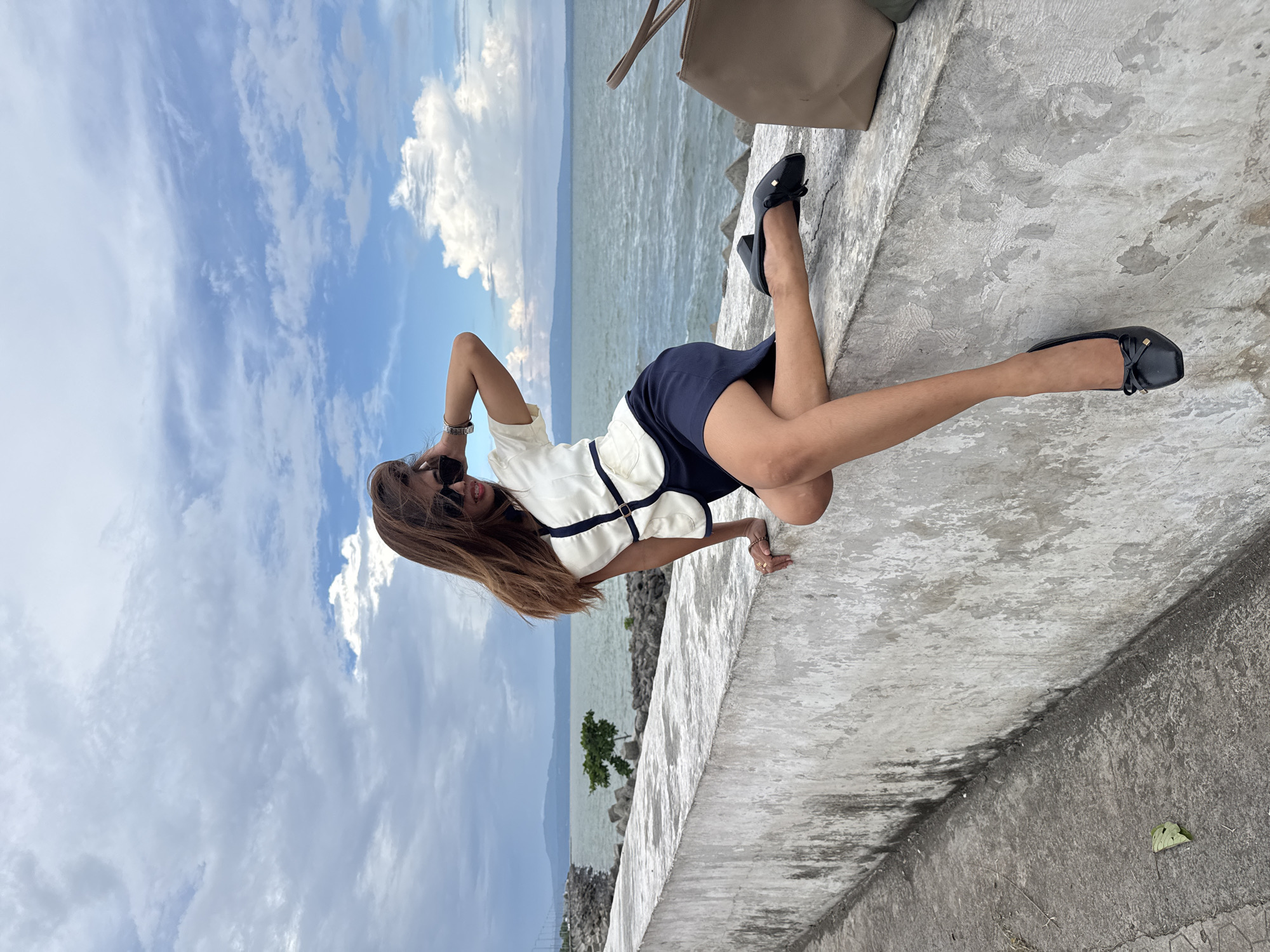 A young woman with long brown hair and sunglasses is sitting on a concrete barrier overlooking a body of water. She is wearing a white and navy blue outfit with black ballet flats. Her right arm is raised and touching her hair, and her left hand is resting on her left knee. The water is calm, and there are mountains in the distance. The sky is cloudy with patches of blue. The concrete barrier is weathered and has some moss growing on it. To the right of the woman is a brown tote bag. The scene is set in Davao City, Philippines, likely along the coast or a body of water within the city limits, possibly near an area with a seawall or promenade.