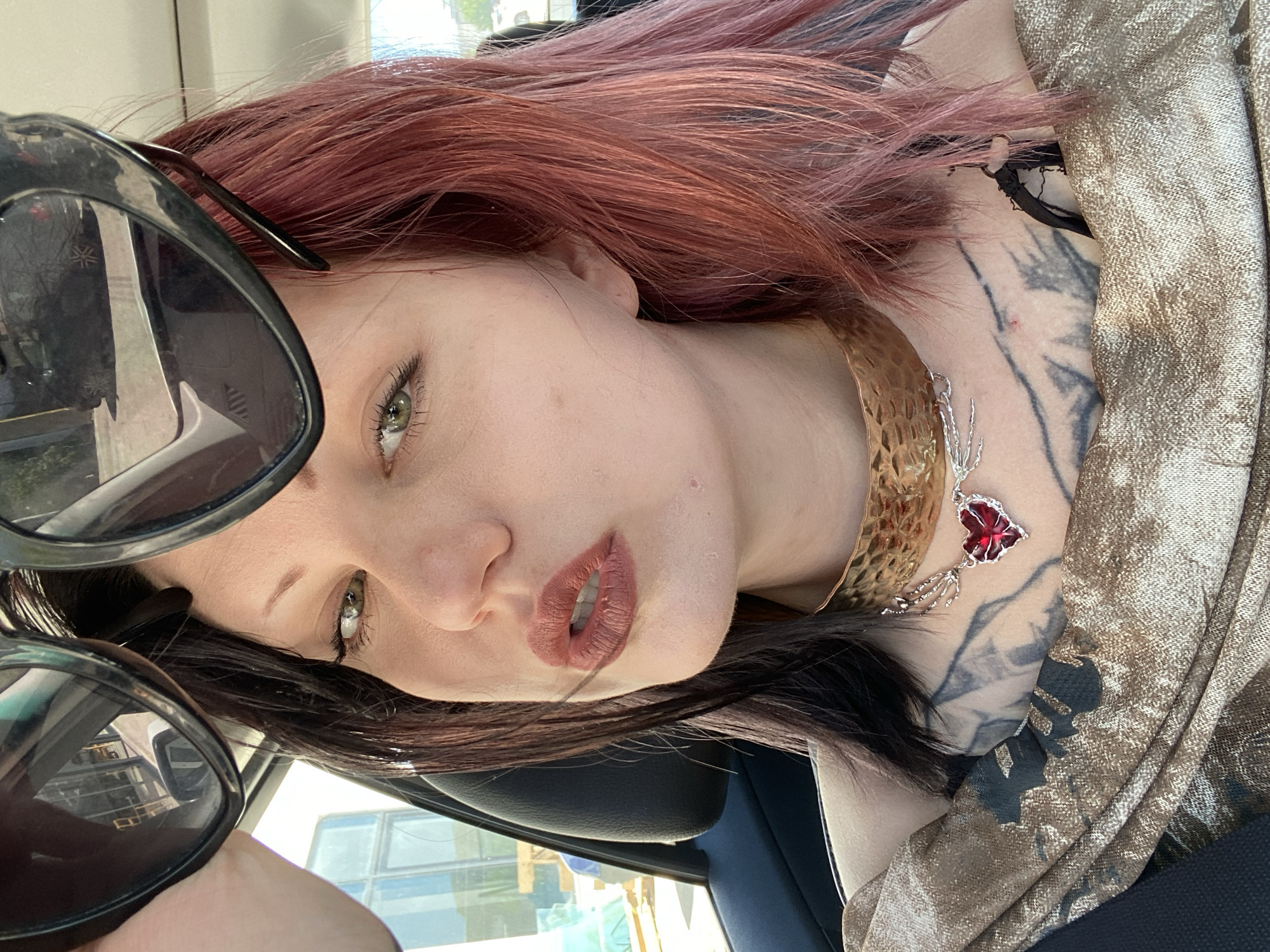 A woman is in a car, wearing a bronze collar necklace with a red stone pendant. She has two-toned hair, dark brown at the roots and a reddish-pink on top. She has fair skin, green eyes, and brown lipstick. A pair of black sunglasses are positioned near her face. A tattoo is visible on her upper chest and neck. The interior of the car has a blue seat and a black headrest. Outside the car window is a building with a white exterior. The city is likely Ulcinj - Ulqin, Montenegro, based on the prompt.