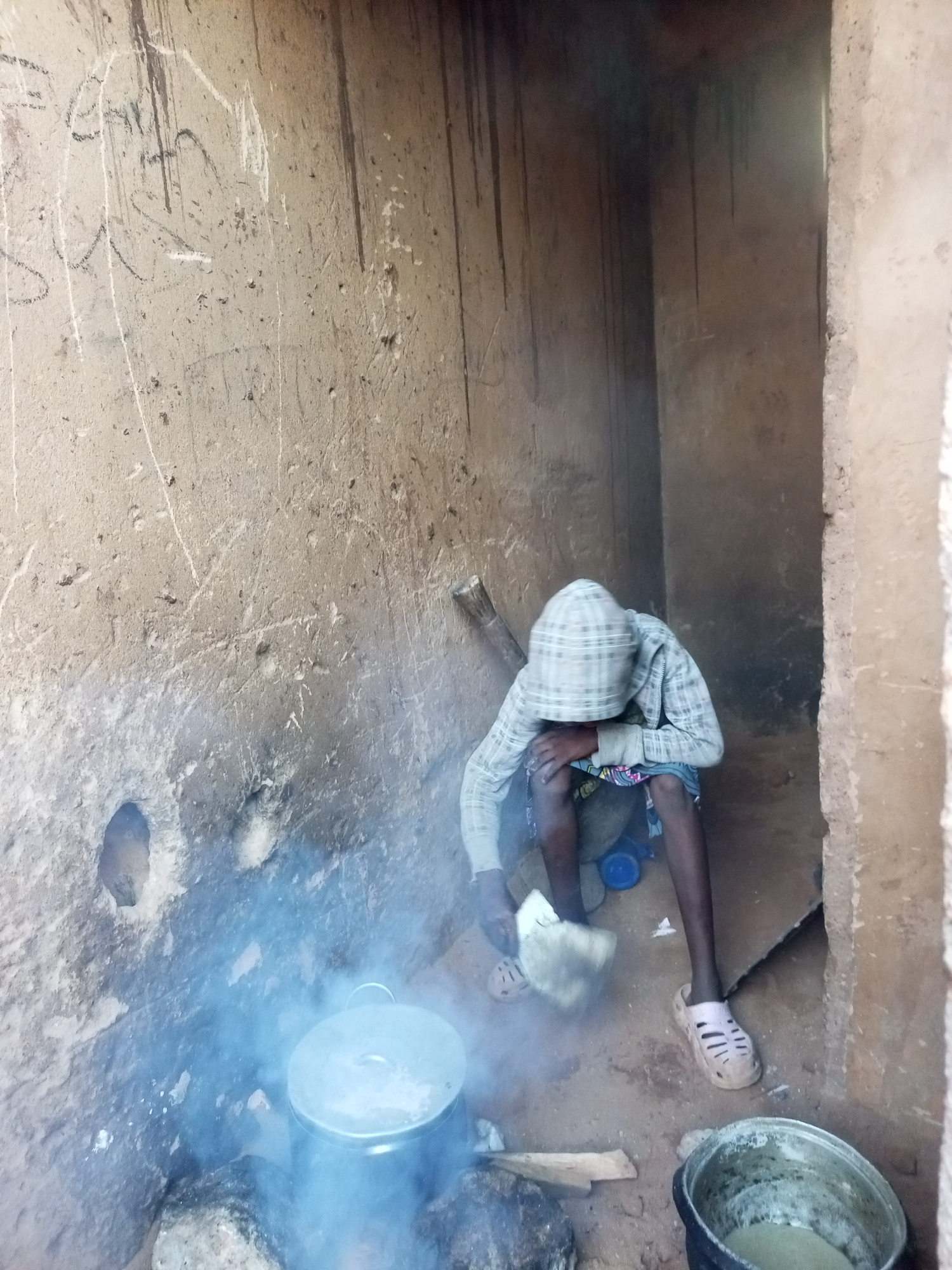 A young person wearing a plaid hooded jacket and a patterned skirt, crouches down in a rough, mud-walled enclosure. The person is tending to a pot cooking over an open fire, indicated by the rising steam. A second pot, containing a light-colored liquid, sits to the right. The enclosure appears to be in an outdoor setting, possibly a rural area in Tempure, Nigeria, given the rustic construction and dry, dusty ground. The walls are marked with lines and scribbles, and there are various debris and stones scattered around the area.