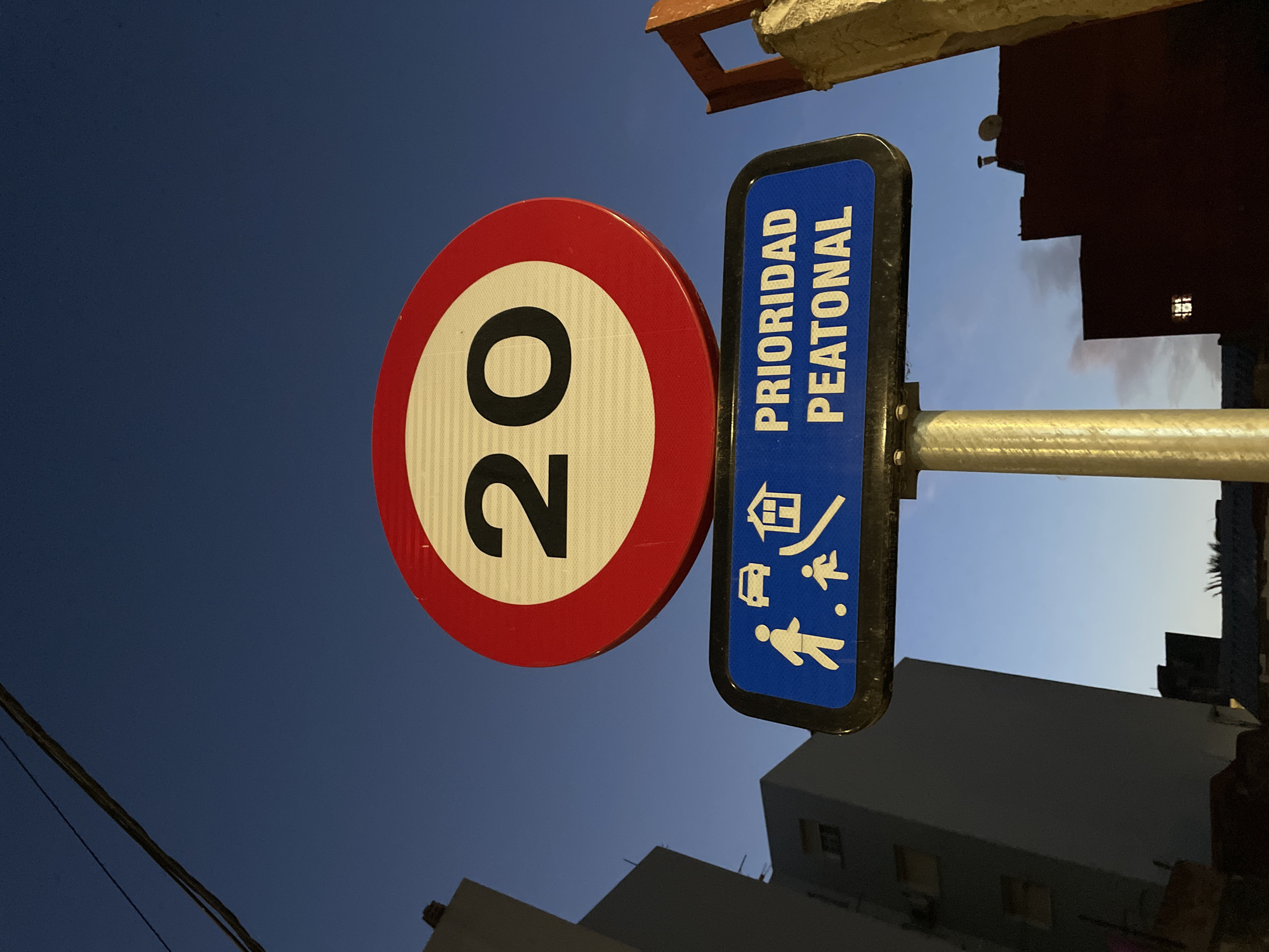 This image captures a street sign in La Línea de la Concepción, Spain, at dusk. The primary subject is a circular speed limit sign displaying "20" within a red border and white background. Below it, a rectangular blue sign reads "PRIORIDAD PEATONAL" (Pedestrian Priority) and features white pictograms of a car, a house with a ramp, and two people, one adult and one child, walking. The signs are mounted on a metal pole. In the background, tall, modern buildings with visible windows are silhouetted against a darkening blue sky. A single power line runs across the top left of the frame. The scene appears to be an urban street setting.