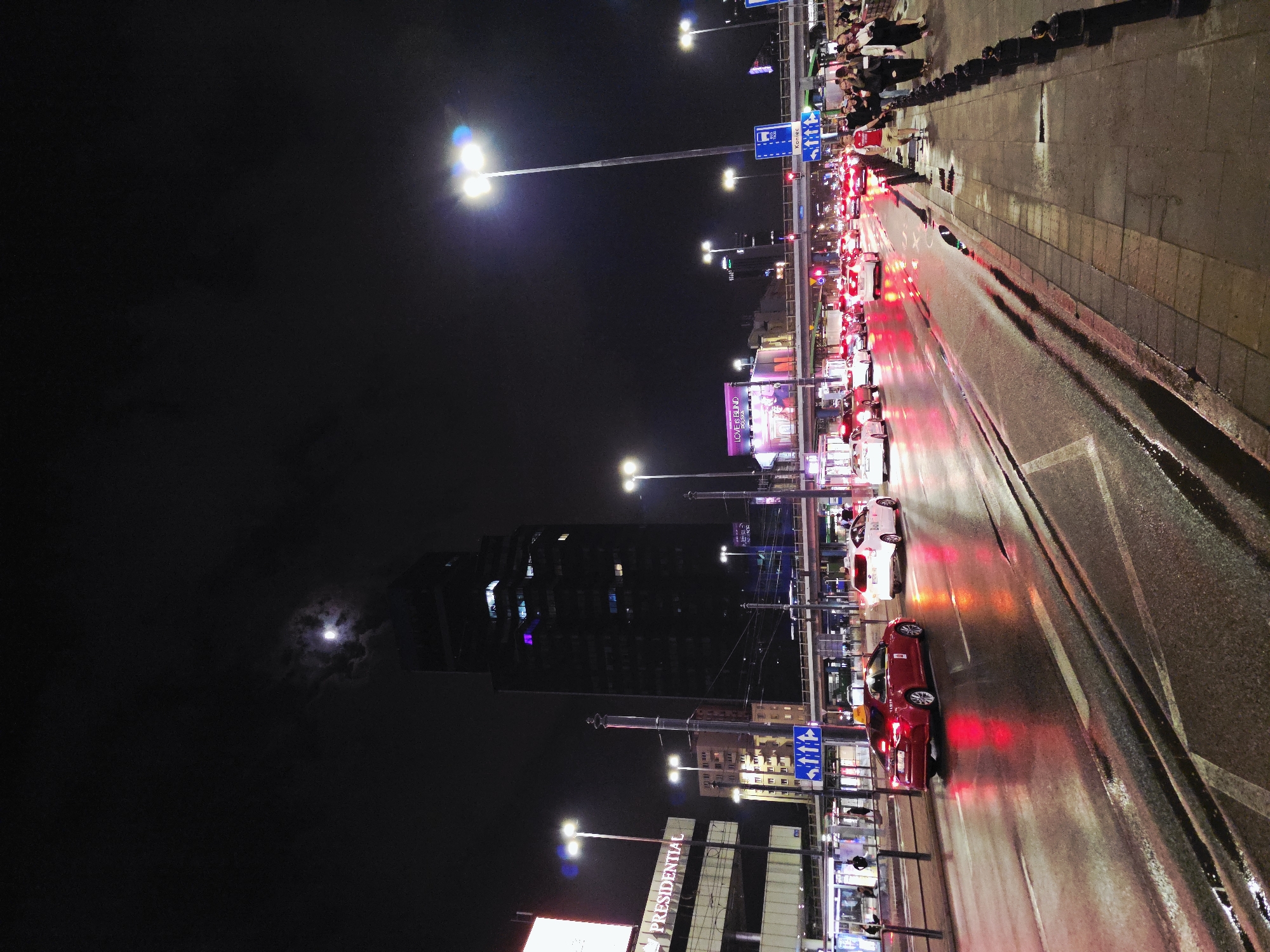 The image depicts a nighttime urban scene in Warsaw, Poland. The view is oriented along a street with moderate traffic, illuminated by streetlights. Red taillights indicate stopped or slow-moving vehicles. A sidewalk runs along the edge of the street where a group of people is standing. In the background, tall buildings are visible, with one prominently displaying the word "PRESIDENTIAL." A billboard reading "LOVE ISLAND" is also visible. The night sky is dark, with a visible moon obscured by clouds. Street signage is present. The environment appears to be wet, with reflections on the road.