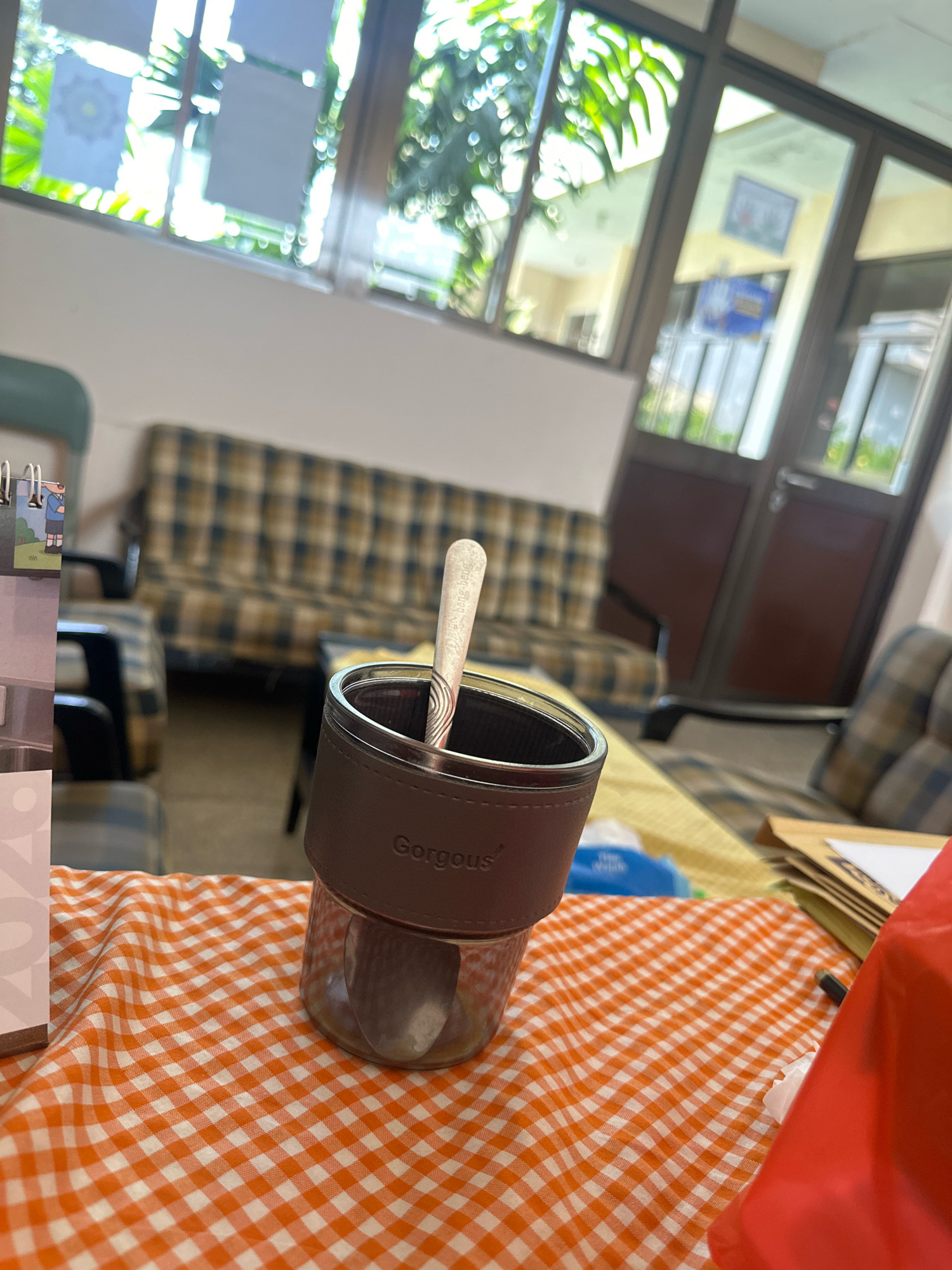 The image captures an indoor scene, possibly an office or a common room, with elements suggesting a mundane daily life setting. A glass jar with a brown sleeve is prominently placed on an orange and white checkered tablecloth. A spoon rests inside the jar, hinting at its use for consumption. In the background, a plaid couch and armchair are visible, along with large windows revealing a blurry outdoor environment with greenery. The overall impression is of a casual, perhaps break time, moment. The image does not contain any specific visual cues to pinpoint its location in Purwotengah, Indonesia, or any other specific city.