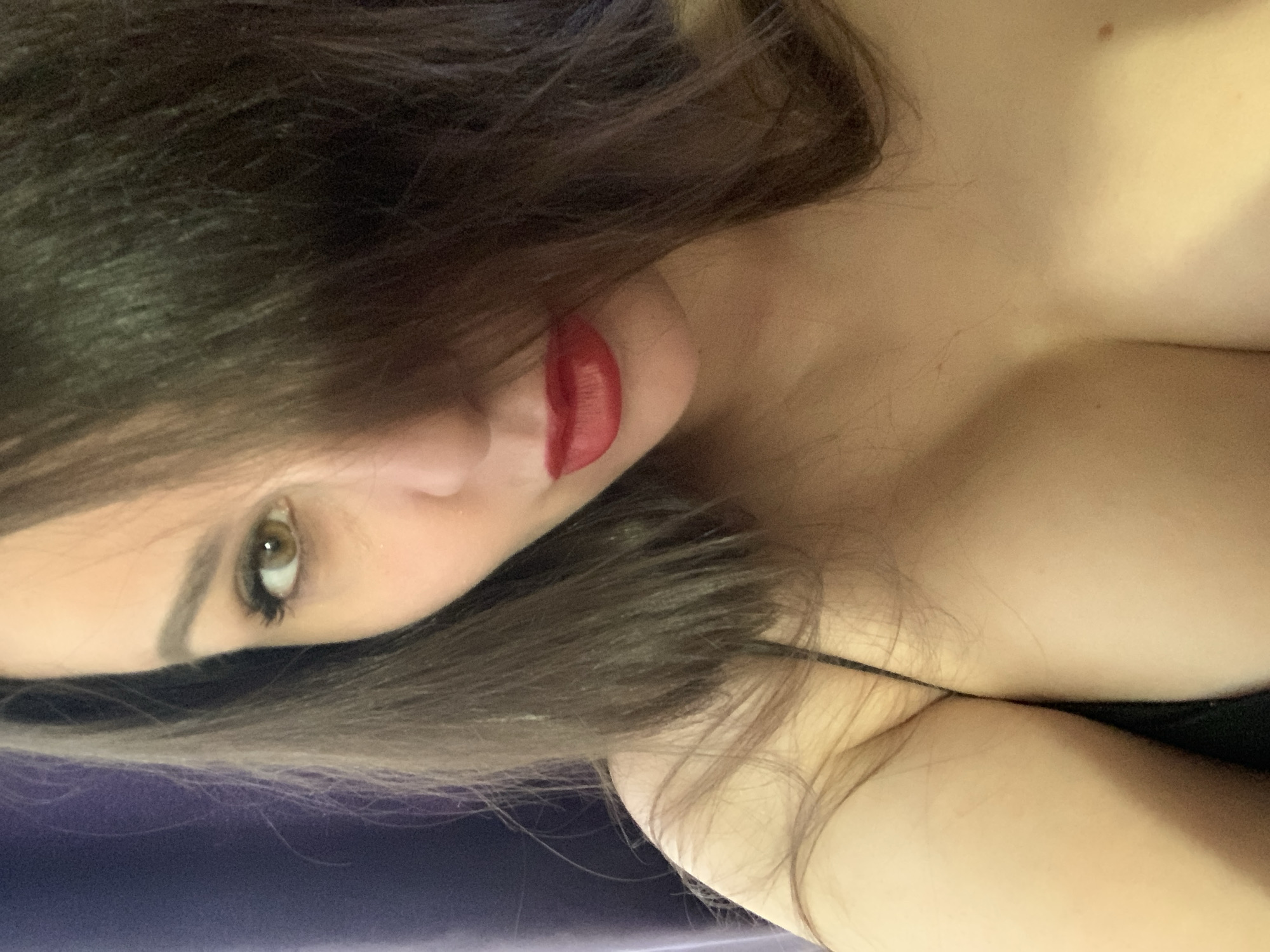 The image features a close-up portrait of an individual, likely female, with dark brown hair partially obscuring the face. The person is looking directly at the camera with a striking olive-green eye. Red lipstick is applied to the lips, which are slightly parted. The visible skin tone is light. The upper body, including a shoulder and the upper chest area, is visible, suggesting a low-cut garment or bare skin. A thin, dark strap is visible across the chest area, possibly from a top or bra. The overall lighting is dim, casting soft shadows, and the background is dark and indistinct, with hints of purple or blue in the lower-left corner. The image is oriented sideways, with the person's head on the left and body extending to the right. No specific activity is taking place other than posing for the photograph. There is no visible text or identifiable objects beyond the person's features and attire. The time of day and specific location cues are unknown.