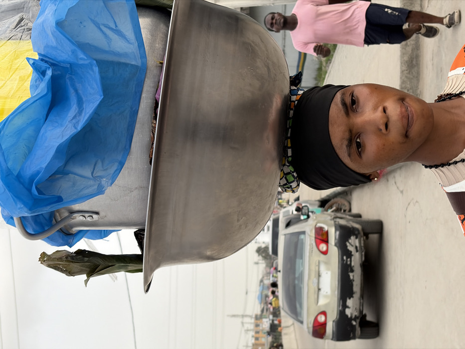 In Jakande, Nigeria, a young individual, likely female, is captured in a close-up, foreground view, carrying a substantial load on her head. She wears a black headwrap over a patterned fabric pad, supporting a large, reflective metallic basin. Stacked within or on top of the basin is a smaller aluminum pot, partially draped with bright blue and yellow plastic sheeting. A dark green, possibly leafy, item protrudes vertically from the contents. She has a subtle nose piercing, a small gold earring, and a dark beaded necklace. Her gaze is directed slightly to the left of the frame, with a neutral expression. In the mid-ground on the right, a male figure in a pink t-shirt and dark shorts walks. On the left, a light-colored sedan with visible paint damage on its rear bumper is parked or moving slowly on a paved road, which extends into the blurred distance with other vehicles and figures under an overcast sky. The scene depicts an urban street environment.