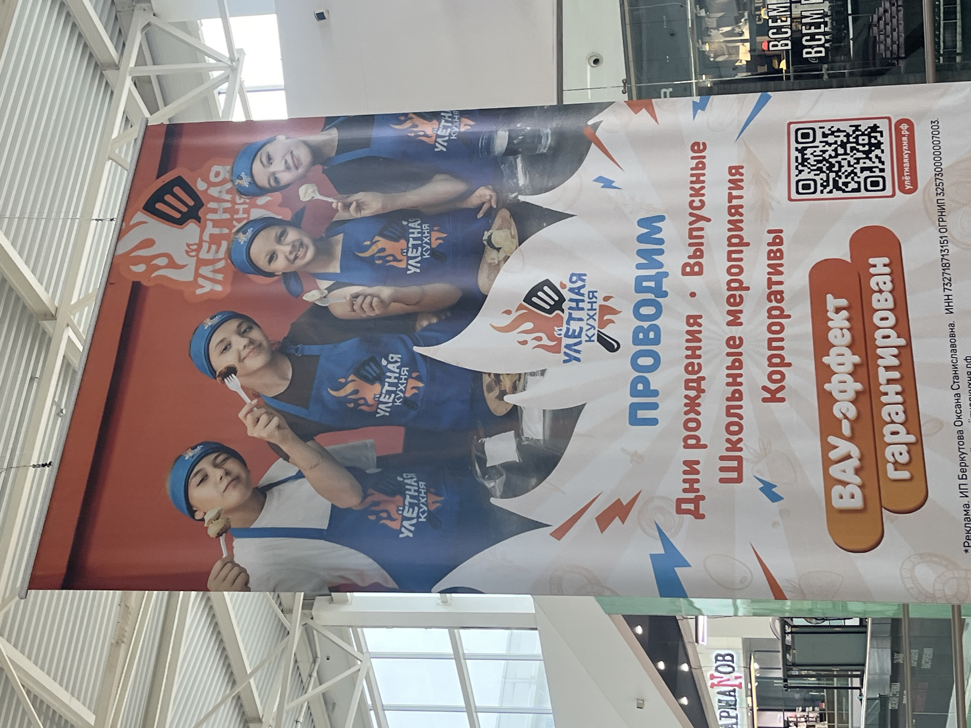 A large vertical banner advertising "УЛЕТНАЯ КУХНЯ" (Awesome Kitchen) is displayed indoors, likely in a commercial building such as a shopping mall or entertainment center, given the high ceiling with exposed metalwork and other background signage. The banner is oriented vertically but appears in the image at a tilted angle.
The main subject of the banner is a group of four young people, appearing to be children or teenagers, smiling and holding spoons or forks with food on them. They are wearing matching blue aprons and headbands, both featuring the "УЛЕТНАЯ КУХНЯ" logo, which depicts a spatula with flames. The overall impression is one of fun and engagement with food preparation or tasting.
The banner's text, primarily in Russian, advertises event hosting services. Key phrases include "ПРОВОДИМ" (We Host) followed by a list of events: "Дни рождения" (Birthdays), "Выпускные" (Graduation parties), "Школьные мероприятия" (School events), and "Корпоративы" (Corporate events). A prominent tagline states "ВАУ-эффект гарантирован" (WOW-effect guaranteed).
A QR code is visible on the banner, leading to the website "улётнаякухня.рф". Below the main text, smaller print reads: "*Реклама. ИП Беркутова Оксана Станиславовна. ИНН 732718713151 ОГРНИП 325730000007003.", indicating that it is an advertisement from an individual entrepreneur.
In the background, hints of other commercial spaces are visible, including partial signs like "АРШАМОВ" and "BCEM". The setting is well-lit, characteristic of an indoor daytime environment. The banner suggests a business in Ulyanovsk, Russia, offering interactive cooking or culinary events for various occasions.