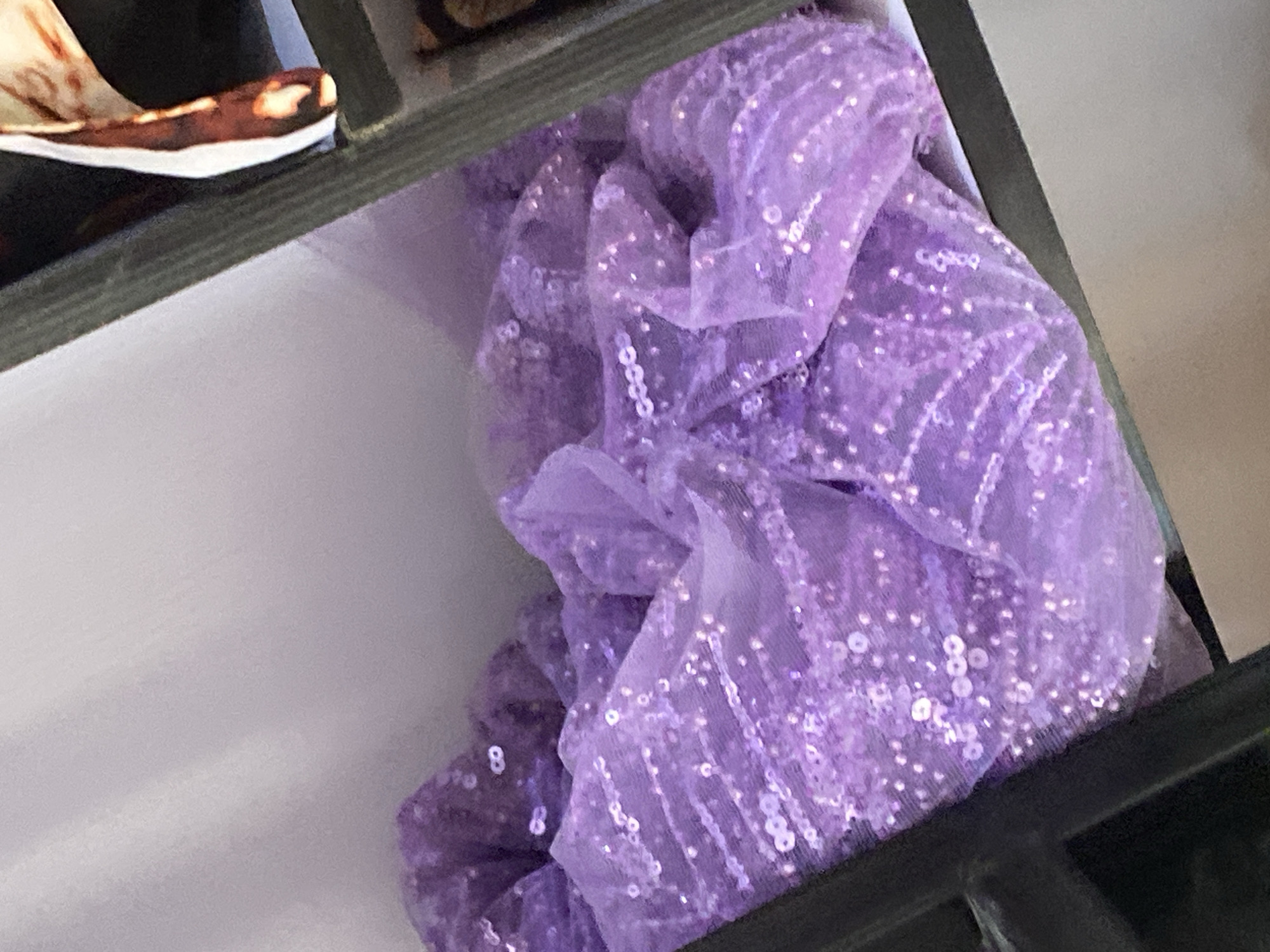 A close-up, slightly blurry, overhead shot captures a swatch of sheer lavender fabric adorned with sparkly sequins. The fabric is draped and bunched, creating soft folds and highlights where the light catches the sequins. The background consists of a pale, muted wall and a dark shelf edge, suggesting the fabric is resting on or within a storage space. No people or animals are visible. The image is devoid of any contextual clues that would place it in Jauro Yinu, Nigeria, or any specific urban environment.