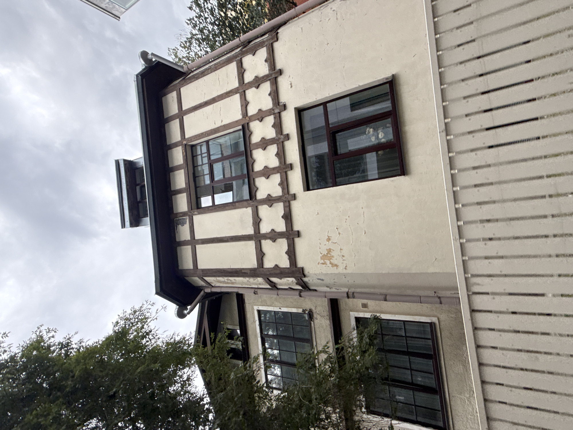 A low-angle shot captures a section of a multi-story building in Tallinn, Estonia, under a cloudy sky. The building features a cream-colored facade with dark brown, timber-frame detailing. Various windows with dark frames are visible, some with reflections showing a white interior. The roofline has dark eaves and a downspout running vertically along the side of the building. To the left, the branches of a dark green tree obscure part of the structure. In the foreground, a white wooden fence runs horizontally across the frame, with vertical slats creating a repetitive pattern. The scene appears to be an exterior view of a residential or commercial property.
