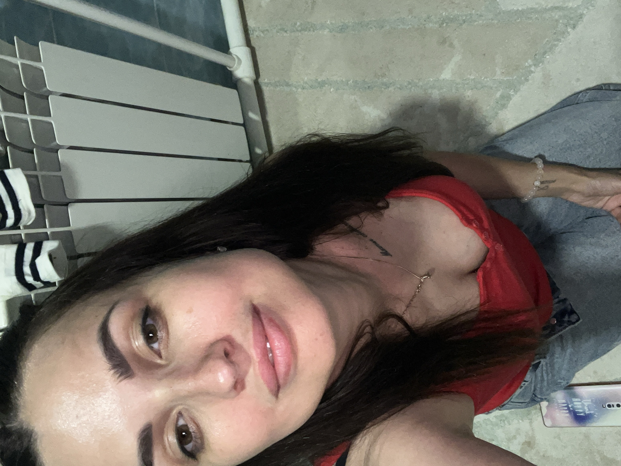 A woman is shown from the chest up in a close-up selfie. She has dark hair, brown eyes, and is wearing a red top and blue jeans. She is kneeling on a floor with a tiled pattern. In the background, a white radiator and a striped cloth are visible.