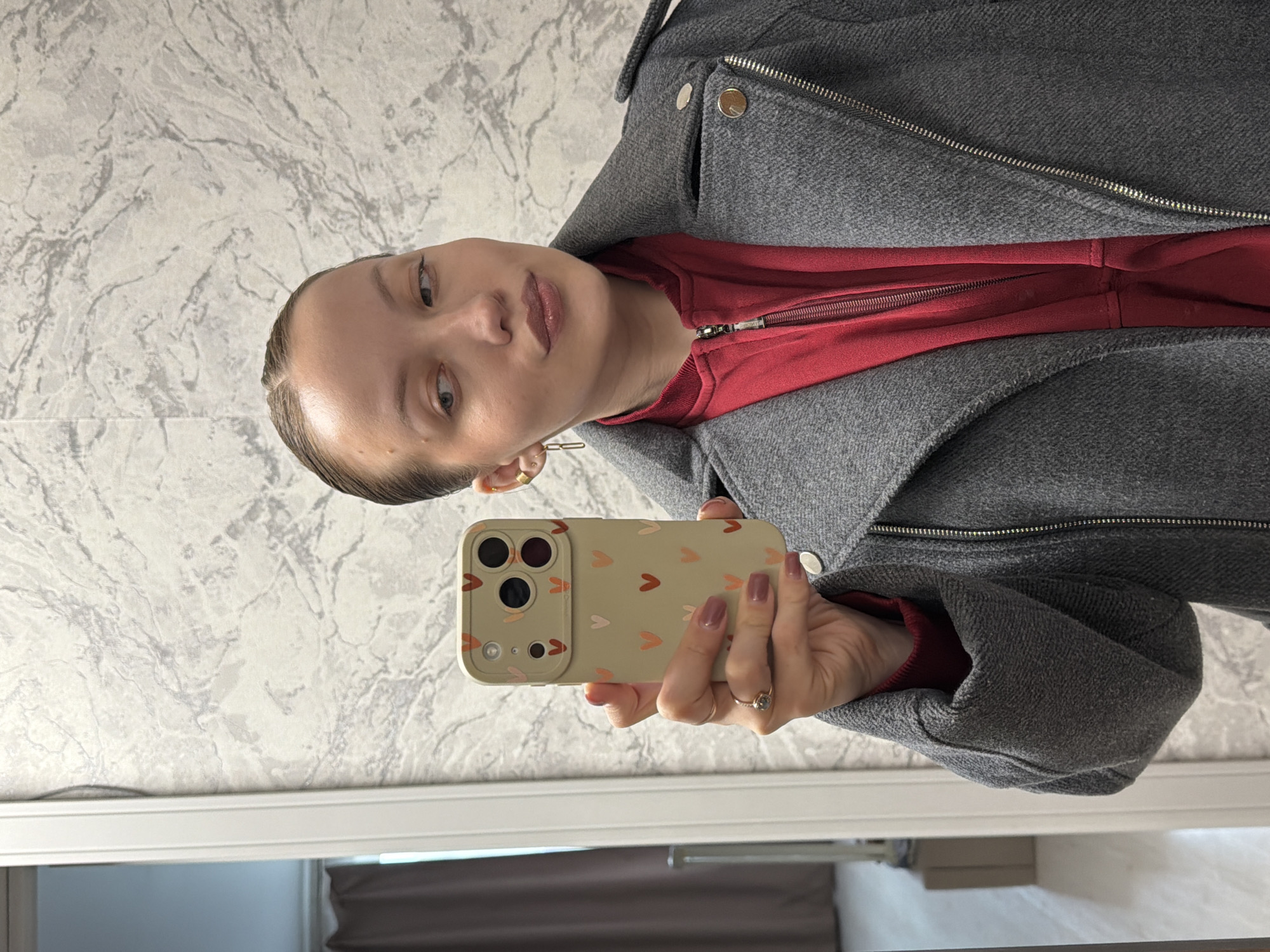 A woman is taking a selfie in a room with a gray and white marble-patterned wall. She is wearing a red hooded sweatshirt and a gray jacket. She is holding a phone with a light-colored case decorated with hearts. The room appears to be indoors, with a white door frame and a glimpse of another room visible in the reflection. The location in Reshetnikovo, Russia, is unknown.