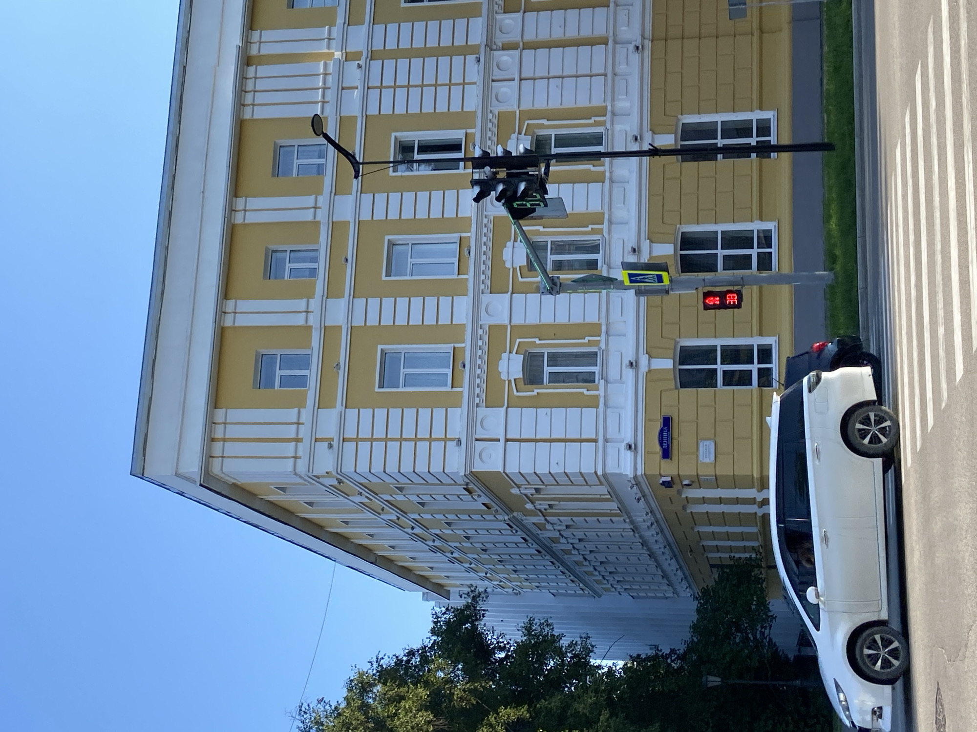 In Blagoveshchensk, Russia, a white minivan is stopped at a pedestrian crossing in front of a yellow building with white trim. The building has numerous windows and decorative architectural elements. A traffic light is visible, showing a red signal and a countdown timer displaying "38". To the left of the traffic light is a sign with a pedestrian symbol. A dark-colored vehicle is partially visible behind the white minivan. Trees are present on the left side of the image, and a clear blue sky is overhead. A street sign with Cyrillic lettering, likely indicating a street name, is affixed to the corner of the yellow building.