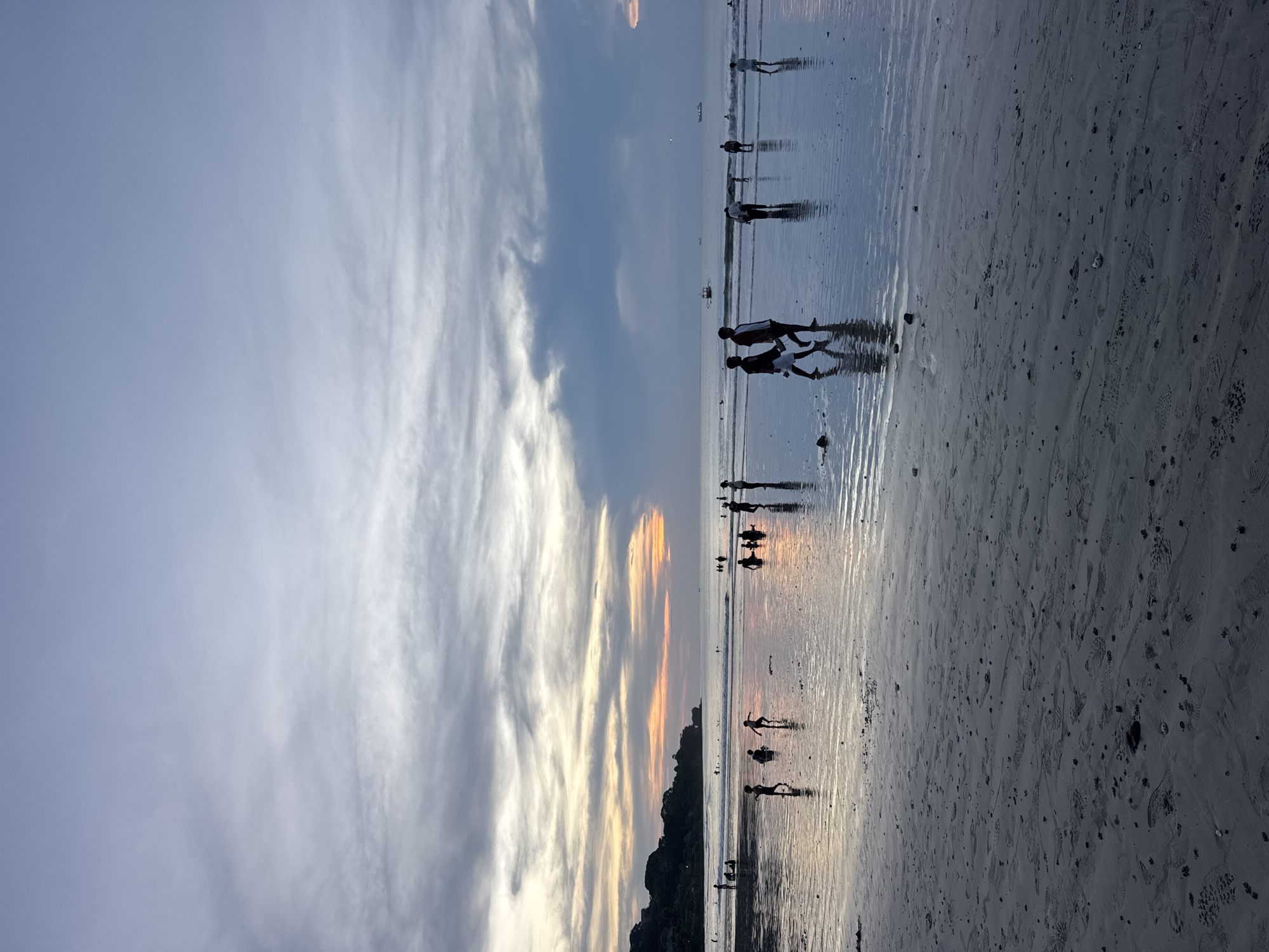 The image depicts a beach at sunset. Several silhouettes of people are visible along the shoreline, with their reflections mirroring in the wet sand. The sky displays a mix of blue and gray clouds, with streaks of orange sunlight piercing through, suggesting the sun is setting near the horizon. A dark landmass can be seen in the lower-left area. The foreground is sandy, with numerous footprints scattered across the surface. The location is in the city of Unknown, Unknown.