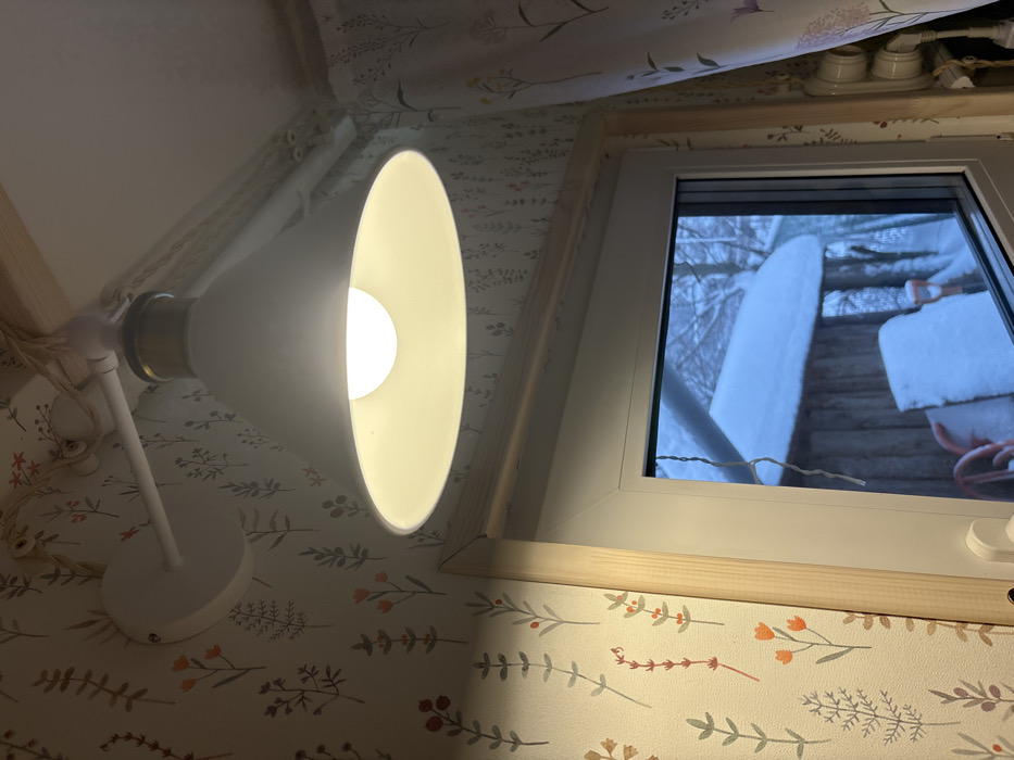The image depicts an interior scene in Lezhnevo, Russia, focused on a window and an overhead light fixture. A white, cone-shaped light fixture with a brass accent ring, mounted on a white arm, illuminates the upper portion of the frame with a bright white bulb. The adjacent wall is covered in light-colored wallpaper featuring a repeating pattern of muted green, orange, and reddish-brown botanical elements. A wooden trim outlines the top and left sides of the white-framed window. Through the window, an exterior winter landscape is visible, characterized by heavy snow covering the ground, a wooden outbuilding with a snow-laden roof, and another snow-covered object, possibly equipment. A thin, clear decorative string hangs vertically inside the window frame. To the far right, a floral curtain with lavender accents and a white electrical outlet with two plugs are partially visible. No individuals are present.