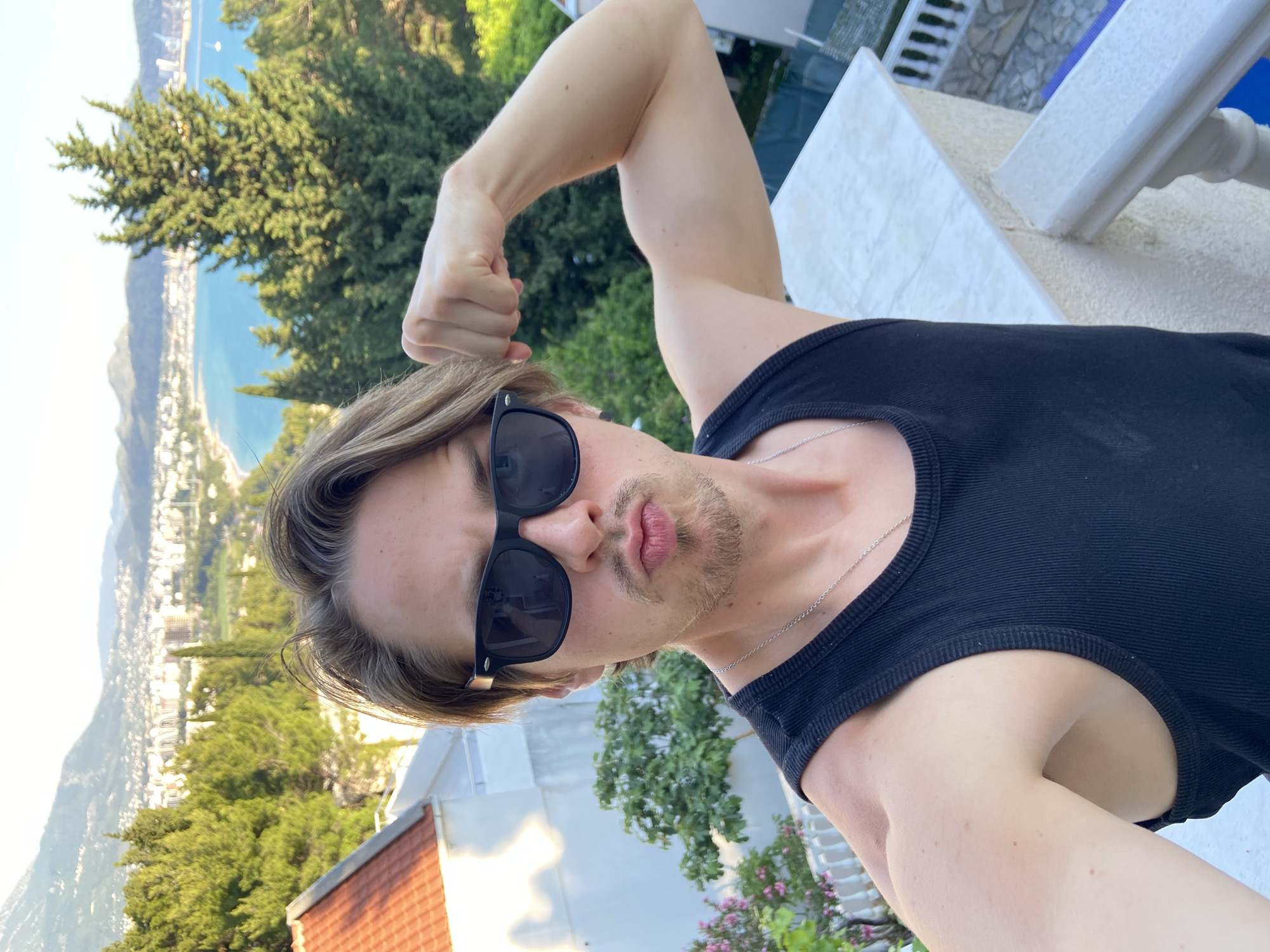 A man in sunglasses and a black tank top poses with his fist clenched, with a view of the coastal city of Šušanj, Montenegro. The city is visible in the background, along with the Adriatic Sea and surrounding mountains. He stands on a balcony with white railings and a partly visible red roof. The location is high enough to provide a panoramic view of the city and sea.
