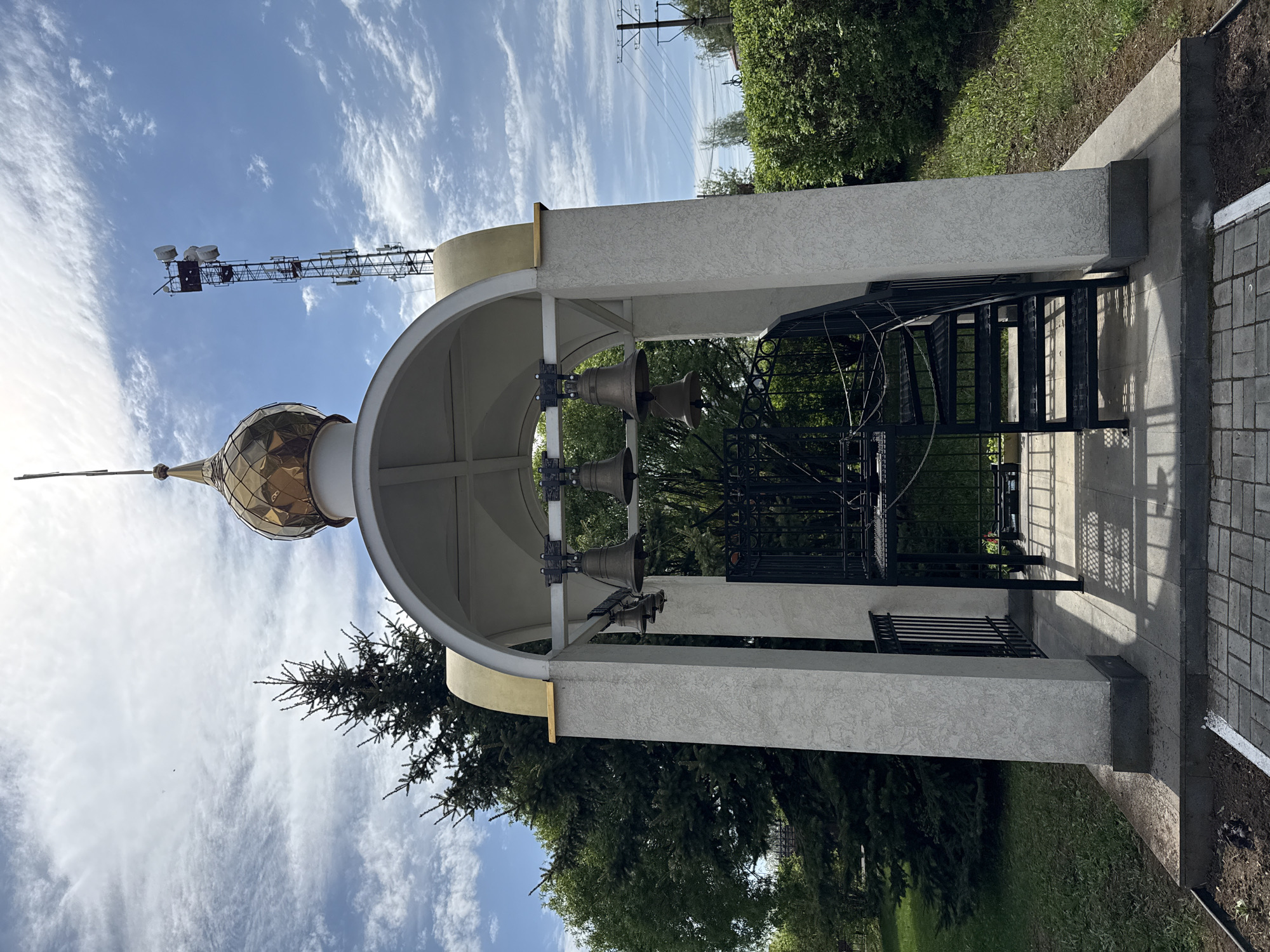 The image shows a bell tower in Votkinsk, Russia, with a golden dome. Three bells are suspended under the arched roof. A black metal fence surrounds the structure. Behind the tower are trees and greenery. The sky is blue with scattered clouds, and a communication tower is visible in the background.