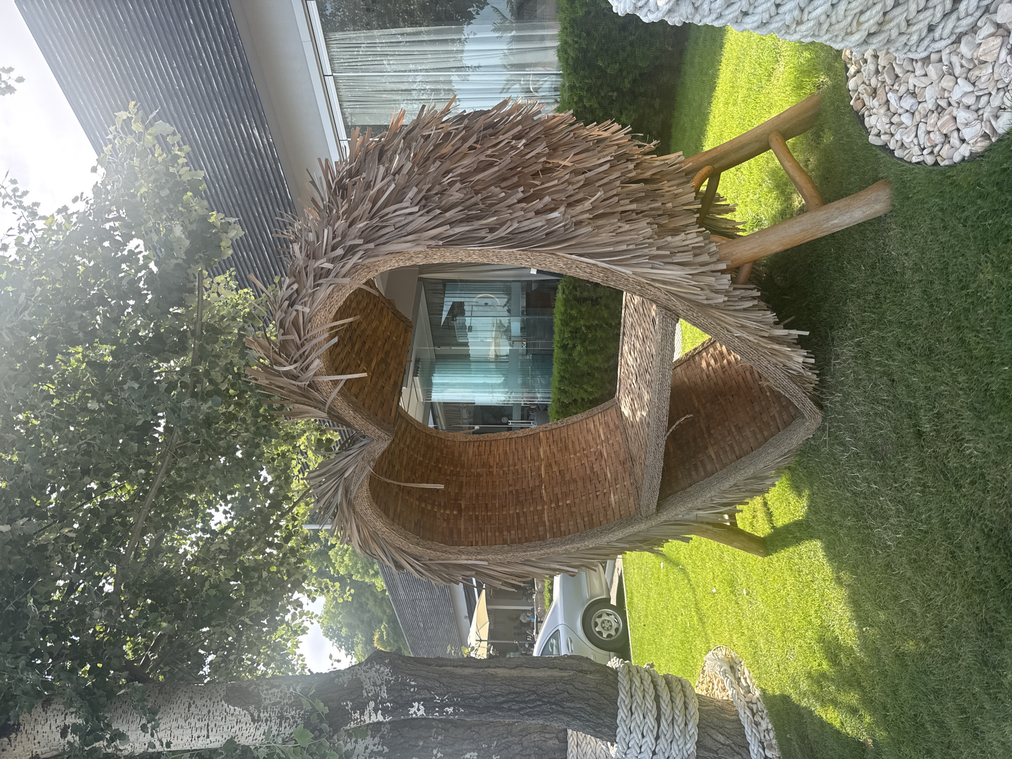 This image captures an outdoor scene in Varna, Bulgaria, on a brightly lit day. The central subject is a large, heart-shaped seating structure crafted from woven natural materials. The inner surface of the heart is composed of tightly woven brown wicker, forming a seat and a backrest. The outer perimeter of the heart is adorned with numerous dried, natural fibers resembling straw or palm fronds, creating a shaggy, textured fringe. The structure is supported by four visible wooden legs, angled for stability.
To the left of the heart structure, a tree with distinctive light-colored, mottled bark stands. Its lower trunk is wrapped with thick, braided white rope in multiple coils. Lush, vibrant green grass covers the ground, illuminated by strong sunlight that casts prominent shadows from the tree and the heart structure. A semi-circular bed of small, light-colored pebbles borders the base of the rope-wrapped tree.
In the mid-ground to the left, partially obscured by the tree, a silver Mercedes-Benz sedan is parked. Beyond the vehicle, an outdoor dining or lounge area is visible with white patio umbrellas and tables, where several indistinct figures are seated. Further back, dense green foliage suggests a wooded or landscaped area.
To the right of the heart structure and in the background, a modern building with a dark, ridged roof is visible. The lower section of the building features large glass windows, reflecting elements of the surrounding environment, including what appears to be light-colored curtains or interior features. A neatly trimmed green hedge runs along the base of the building. The sunlight originates from the upper right, casting a bright glare and highlighting the upper foliage of trees. The sky is partially visible, appearing bright and clear.