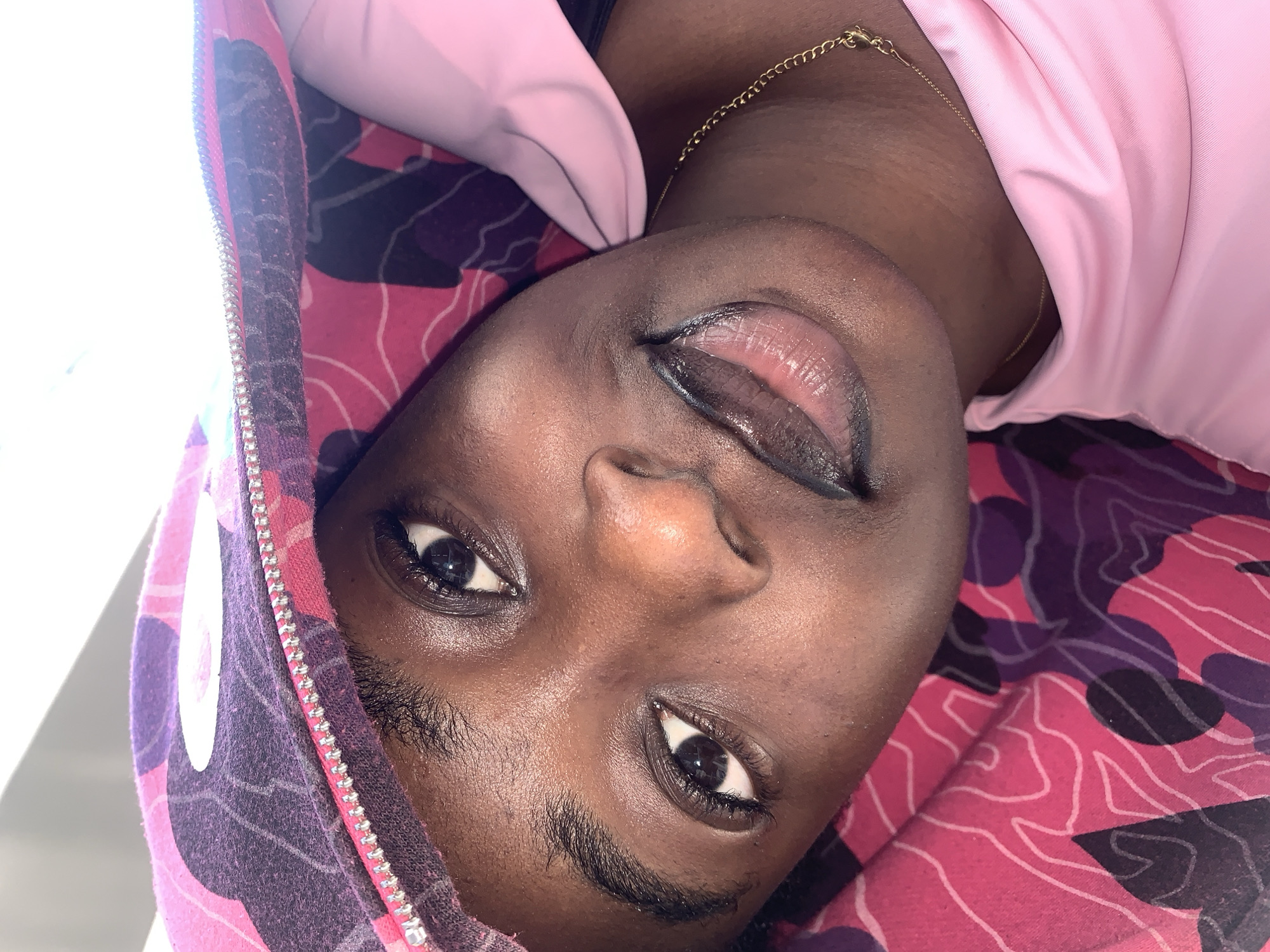 A close-up, head-and-shoulders shot features a young Black woman lying down, looking upwards. She is wearing a pink top and a colorful hoodie with a pink and purple camouflage pattern, which is pulled up over her head, partially obscuring her face. The woman has dark eyes, dark eyebrows, and full lips with a glossy pink lip gloss. A thin gold chain necklace is visible around her neck. The background appears to be an indoor setting with bright, white lighting coming from above. The image does not provide any specific details or context that would allow for the determination of a location in Gura Top, Nigeria.