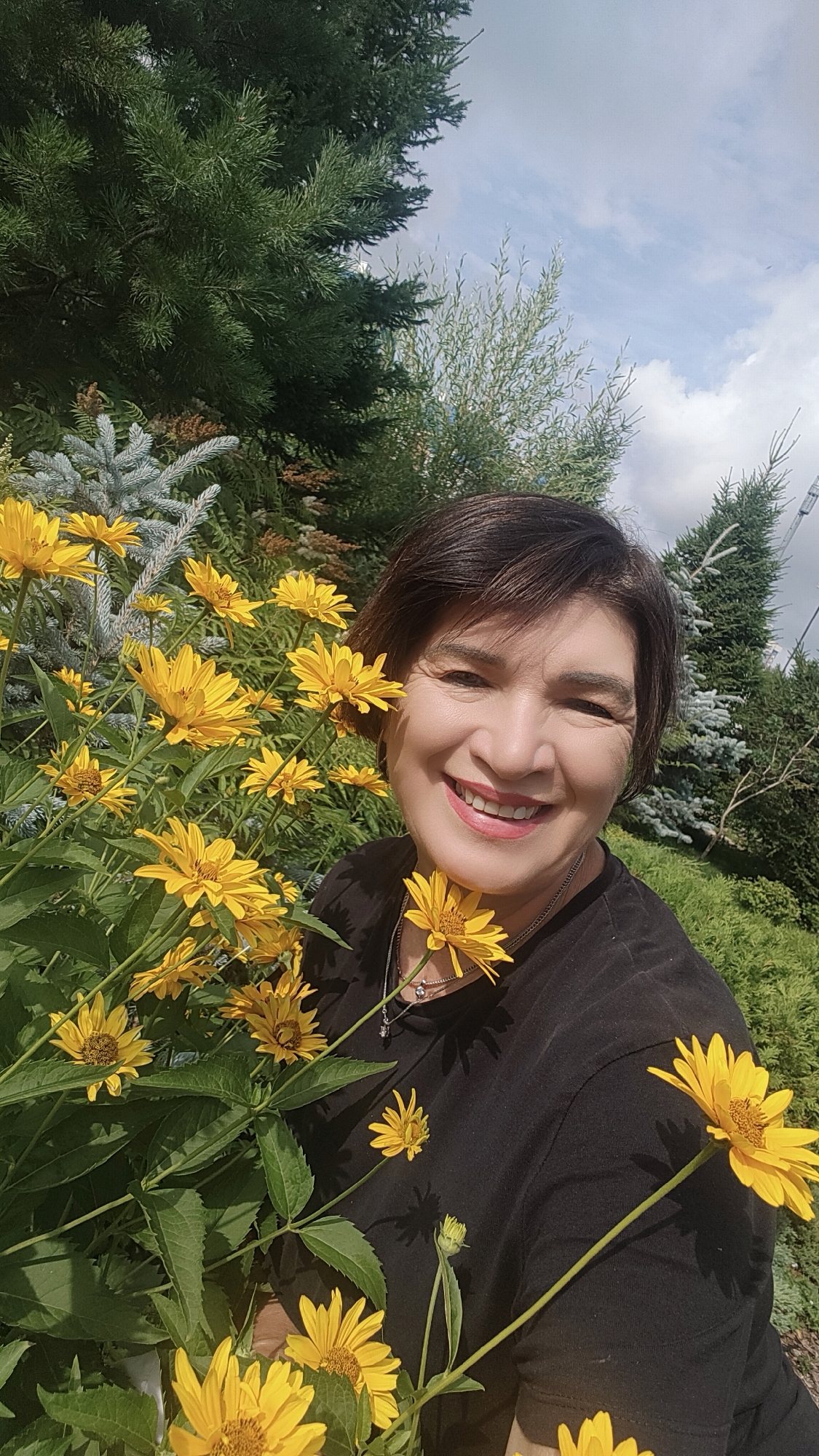 A woman with short brown hair smiles at the camera, positioned to the right of a dense cluster of yellow flowers. She is wearing a black t-shirt and a silver necklace. The background consists of green trees and a cloudy sky. The image appears to be a selfie taken outdoors in a garden or park setting. The specific location within the city of Krasnoyarsk, Russia, is not discernible from the visual information alone.