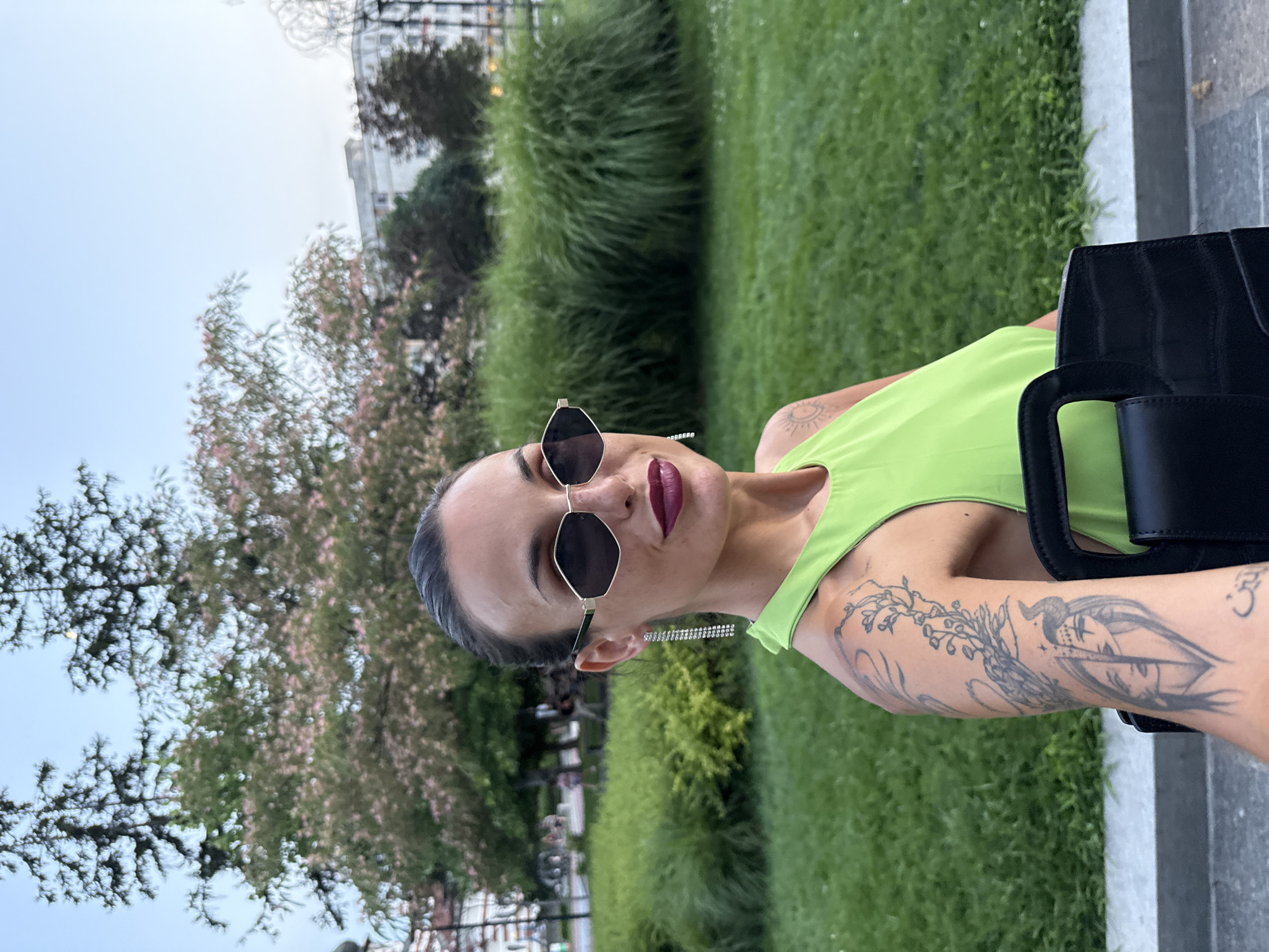 In the city of Pleven, Bulgaria, a woman poses for a photo outdoors. She is wearing a lime green halter top, large angular sunglasses, and dangling diamond earrings. Her dark hair is pulled back into a bun. She has a tattoo on her left arm. In the background, there is a green lawn, tall grass, trees with pink flowers, and a building. The woman is holding a black handbag.