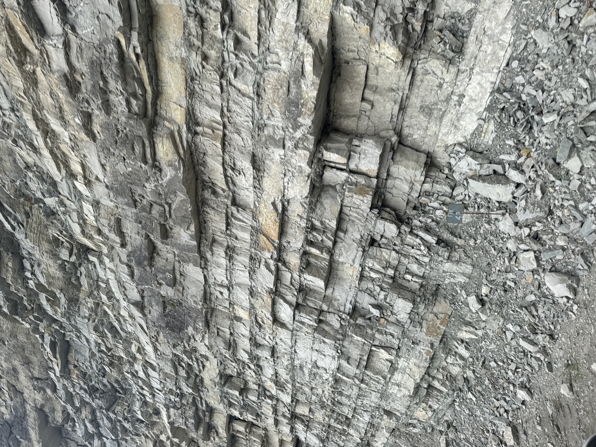 The image shows a steep rock face with distinct horizontal striations, indicating layers of sedimentary rock. The rock is predominantly gray, with some lighter, yellowish-brown layers. The rock face is fractured and uneven, with many loose stones and debris at its base. There is a small signpost in the lower right quadrant of the image, partially obscured by rocks, which appears to have some markings on it. The environment appears to be a natural landscape, likely a roadside cut or cliffside. There are no people or animals visible in the image.