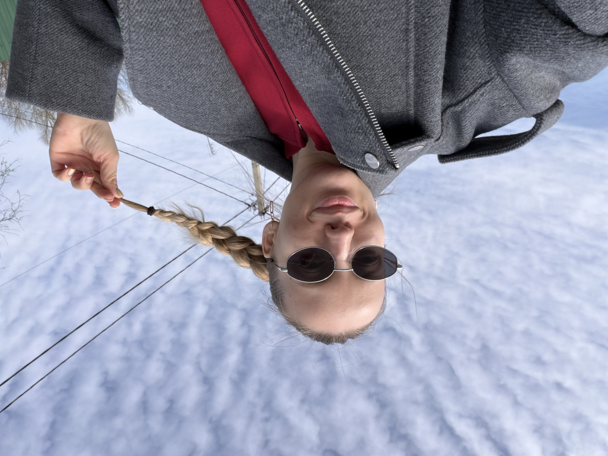The image shows a woman in a grey jacket over a red top, wearing sunglasses, with her blonde hair in a braid. She is holding the end of her braid. The background consists of a cloudy blue sky, overhead power lines, and bare tree branches. The image appears to have been taken from a low angle, facing upwards towards the woman. The location is unspecified, and there is no indication the image was taken in Reshetnikovo, Russia.