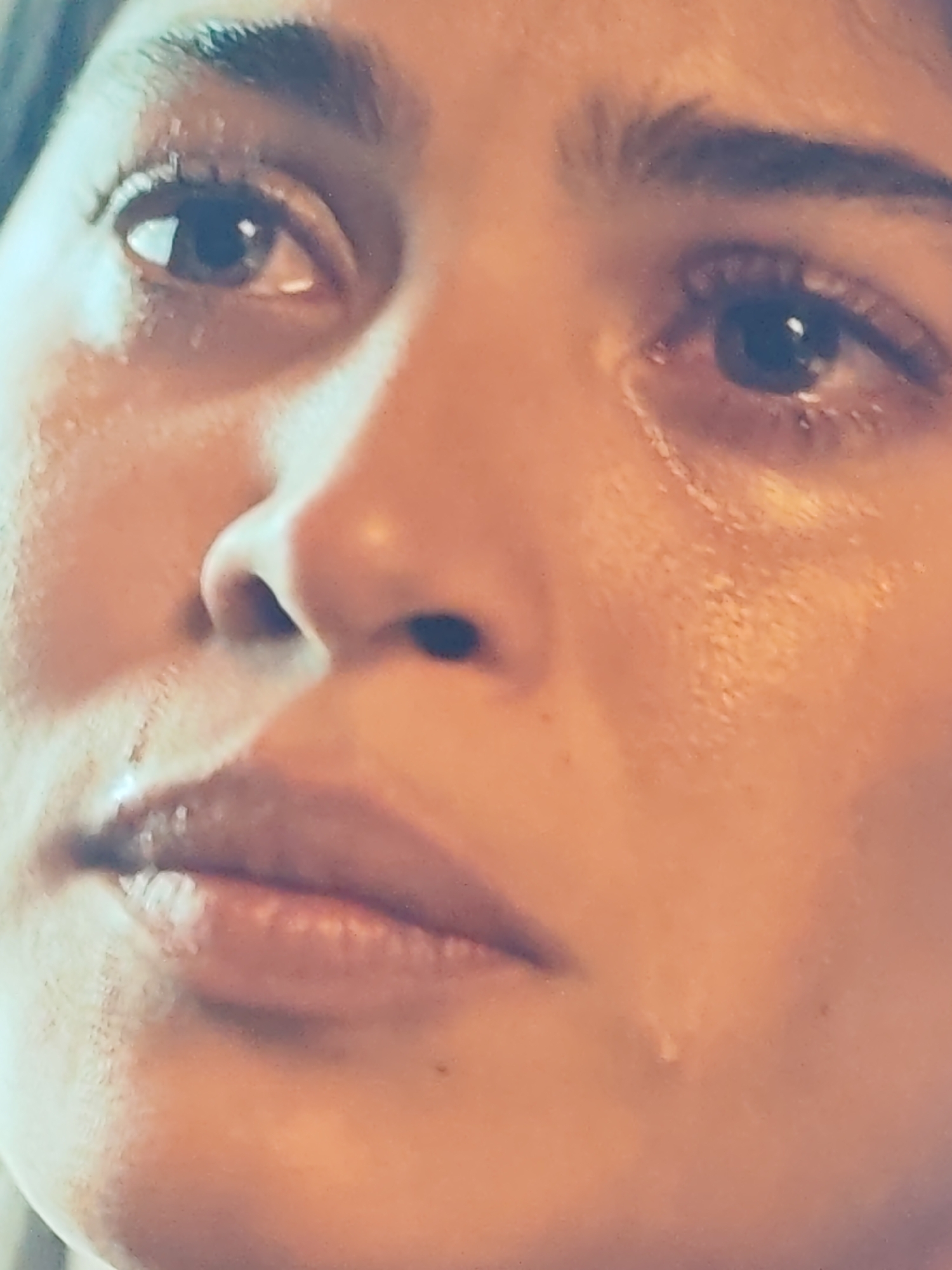 A close-up, head-and-shoulders shot shows a person's face, with a focus on their eyes, nose, and mouth. The person's skin appears wet, with visible tears running down their cheeks and pooling around their eyes. The lighting is soft and warm, casting a gentle glow on their features. The background is out of focus, making it difficult to determine the setting. The person's expression is one of sadness or distress, with their eyes downcast and their lips slightly parted.
