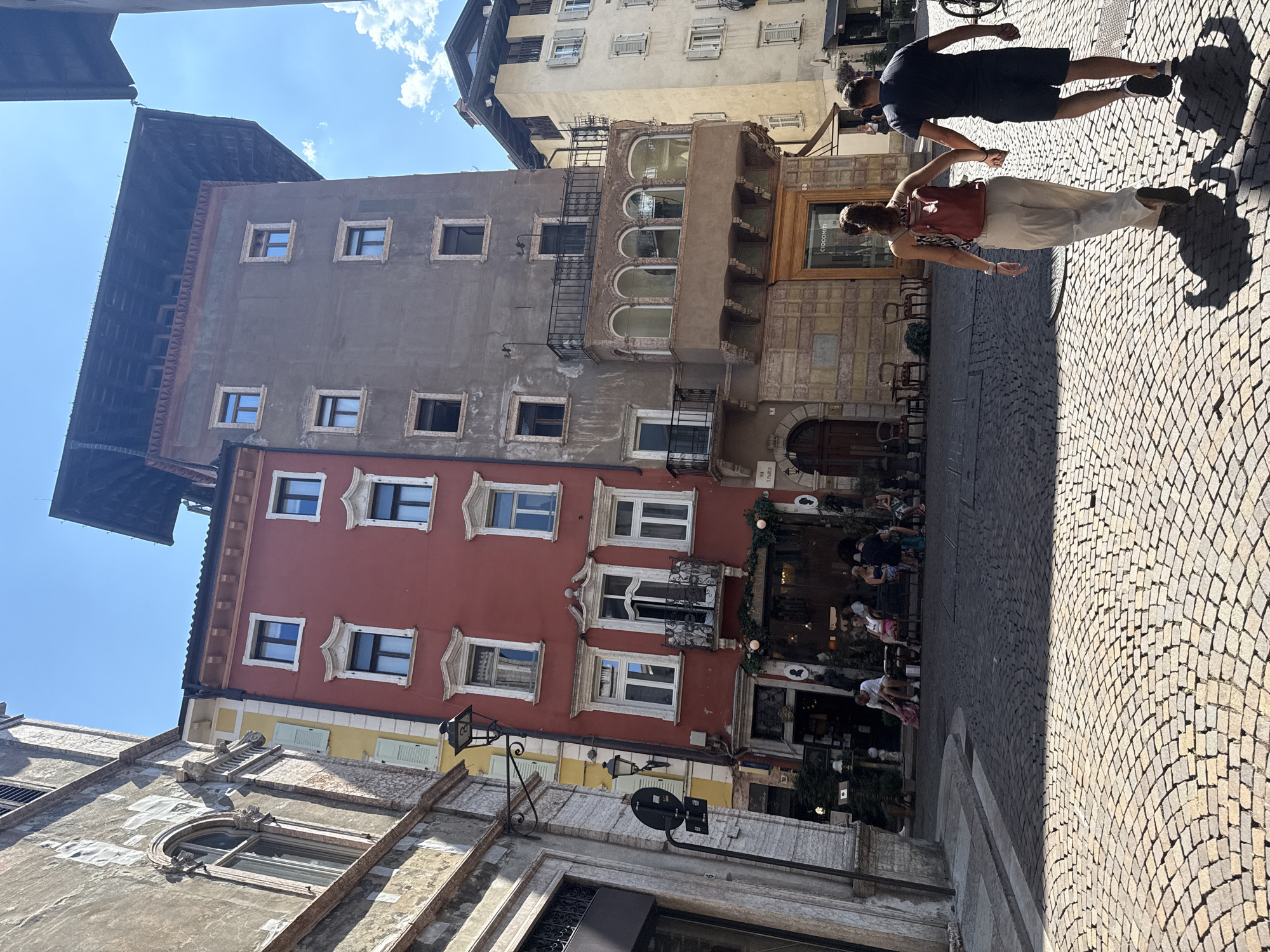 The image shows a street scene in Trento, Italy, with several multi-story buildings lining a cobblestone street. In the foreground, a couple is walking away from the camera, hand in hand. The woman is wearing a red backpack and light-colored pants, and the man is wearing a dark t-shirt and shorts. Both are casting long shadows on the cobblestones, indicating a sunny day.
The buildings have a mix of architectural styles and colors. The building on the left is a light-colored stone structure with arched windows and decorative elements. Adjacent to it is a vibrant red building with white-trimmed windows and a balcony on the second floor. To the right of the red building is a taller, grey building with a distinctive dark, overhanging roof structure. This building has several windows and a unique enclosed balcony with arched windows on one level. Further down the street, a lighter-colored building is visible.
Several people are seated at outdoor tables in front of a cafe or restaurant, suggesting a leisurely atmosphere. Signs and awnings are visible on the ground floor of the buildings. The cobblestone pavement is well-maintained and curves through the street. The sky is blue with some scattered white clouds. The scene depicts a typical European street with historical architecture and pedestrian activity.