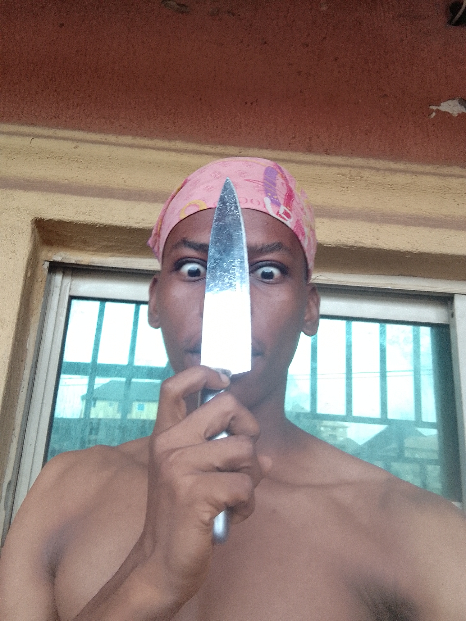 A shirtless person wearing a pink headscarf holds a large kitchen knife in front of their face, obscuring their nose. The eyes are wide, suggesting surprise or shock. The person stands in front of a window. Reflected in the window are buildings, indicating an urban environment. The setting appears to be Nkpor, Nigeria. A building wall with an orange-painted upper section and a yellow-painted lower section is visible above the window.