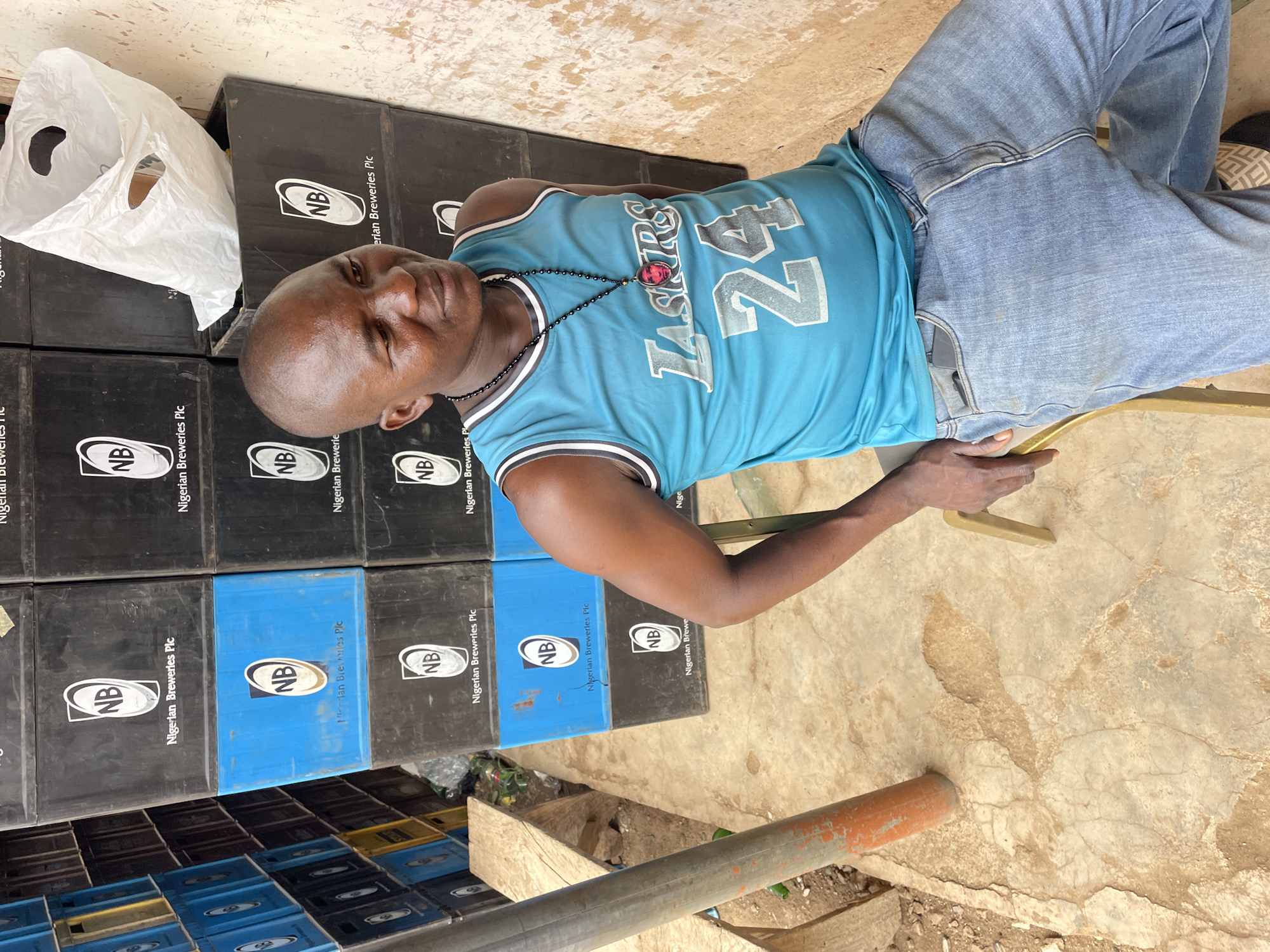 A man wearing a blue "Lakers 24" jersey and jeans sits on a chair next to a stack of beer crates with the "Nigerian Breweries Plc" logo. A white plastic bag sits atop the crates. The setting appears to be outdoors, with a rough, tiled or concrete ground surface. The photograph was likely taken in or near Bukuru, Nigeria due to the presence of the "Nigerian Breweries Plc" crates.