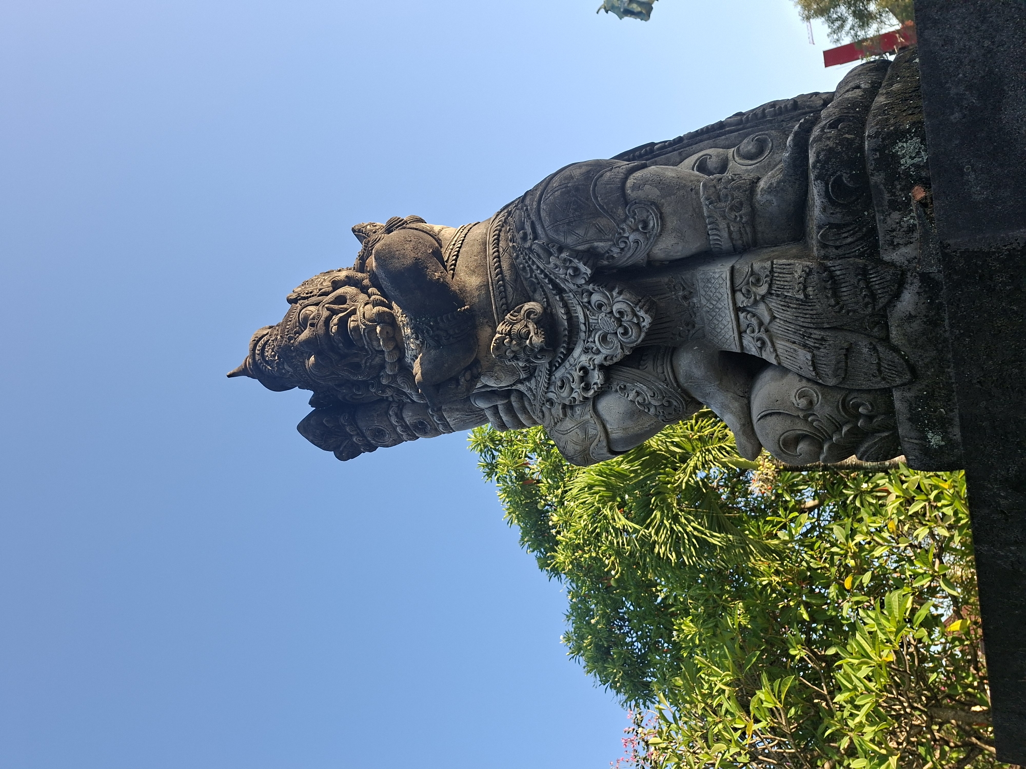 A low-angle, full shot captures a large, stone statue of a demonic figure against a clear blue sky. The statue is carved with intricate details, featuring a fierce expression, elaborate headdress, and ornate clothing. To the left of the statue, lush green trees with vibrant foliage fill the background, with hints of pink flowers visible. The statue is positioned on a dark, rough-textured pedestal, and a small patch of moss and a fallen leaf are visible on its surface. In the far right background, a distant red structure and a construction crane suggest an urban or developed area, potentially the city of Gianyar, Indonesia. The sunlight casts subtle shadows on the statue, highlighting its weathered texture.