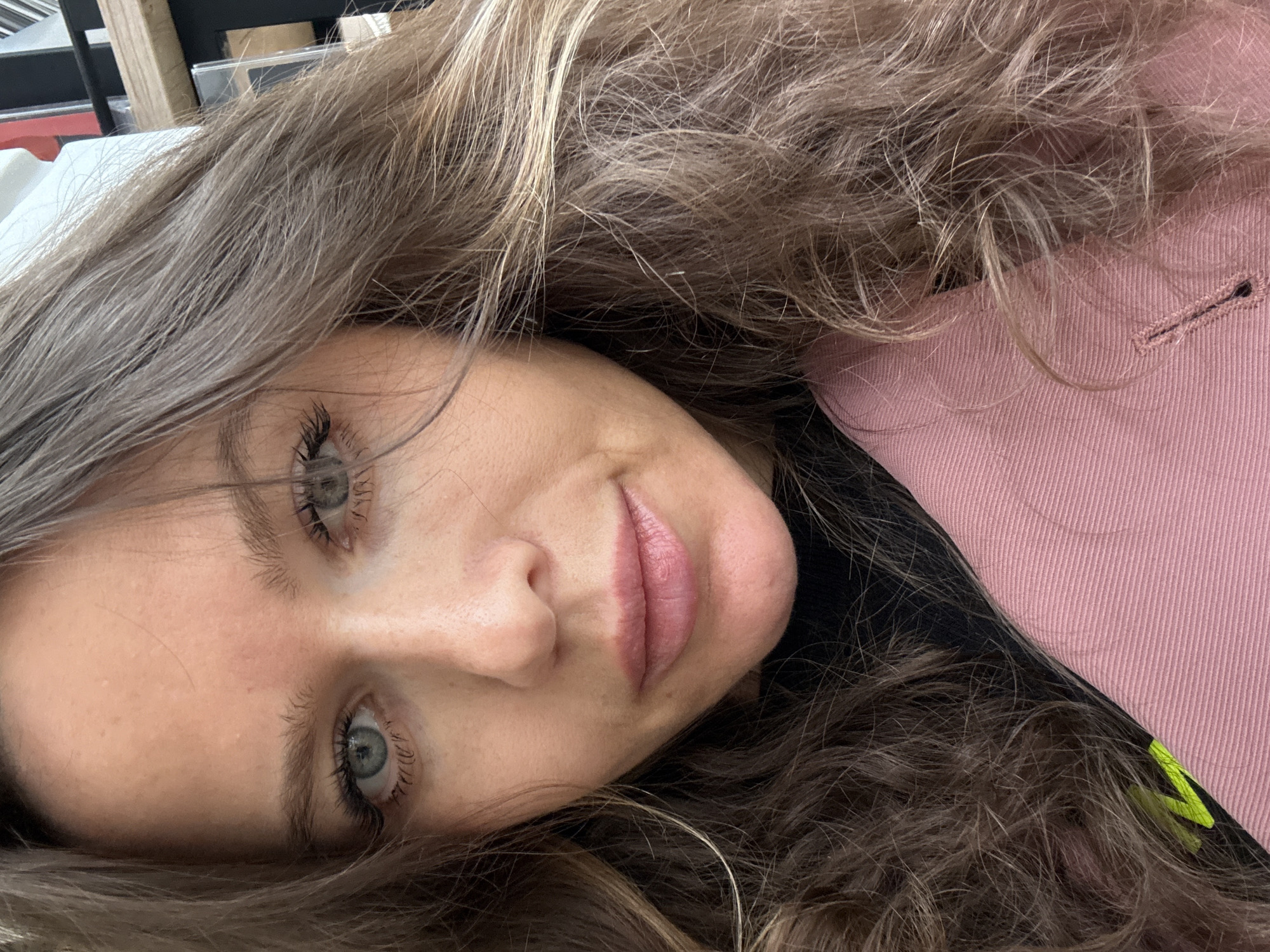 This is a close-up portrait of a young woman, likely a selfie, taken in Moscow, Russia. Her head is tilted, giving the impression she is lying down or leaning back. She has long, wavy, light brown hair with blonde highlights that frames her face and spreads around her.
Her face is the central focus, illuminated by soft, natural-looking light. She has light-colored eyes (appearing blue or grey-green), enhanced by dark mascara on her eyelashes. Her eyebrows are naturally shaped, and her fair skin shows a few subtle freckles on her forehead and cheeks. Her lips are a natural pink with a slight rosy tint, forming a gentle, soft smile.
She is wearing a pink garment, possibly a jacket or a thick shirt, visible on the right side of the frame. The fabric appears to be textured, with a visible buttonhole detail. Underneath the pink garment, a darker, possibly black, fabric is visible. A small, bright yellow-green detail, perhaps a strap or a tag, peeks out near her hair on the lower right side.
The background is softly blurred and indistinct, suggesting an indoor setting with muted tones, possibly showing shelves or furniture. The overall impression is one of calm and relaxation, with a subtle, friendly expression. No specific activities or events are depicted beyond the portrait itself. No visible text is present in the image.