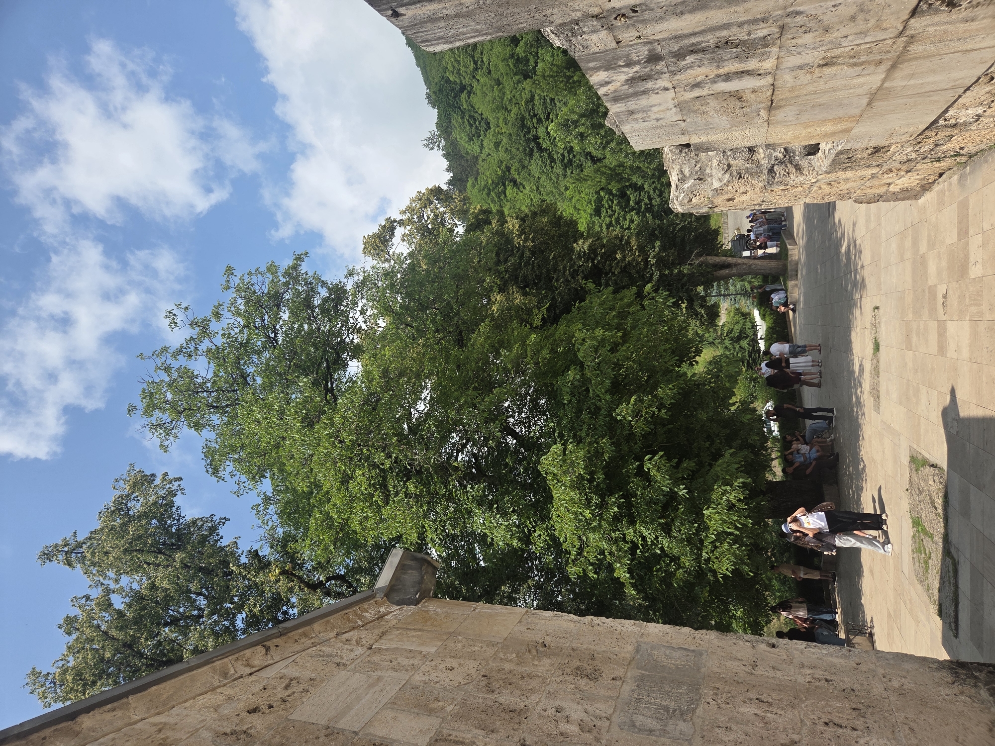 In Teghut, Armenia, a sunny day unfolds at an ancient site, where stone walls of rough-hewn blocks frame a courtyard paved with square flagstones. Lush green trees, including a large specimen prominently in the foreground, cast dappled shadows. In the distance, a verdant hillside cloaked in dense forest rises under a bright blue sky dotted with white clouds. A group of tourists are scattered throughout the courtyard. Several individuals are standing or sitting near the base of the large tree, while others are seen walking along the paved path. Two women in the foreground are posing for a photo, with one capturing the image. Other people are visible in the background, observing the scenery or conversing. The scene conveys a sense of historical exploration and leisurely tourism against a backdrop of natural beauty.