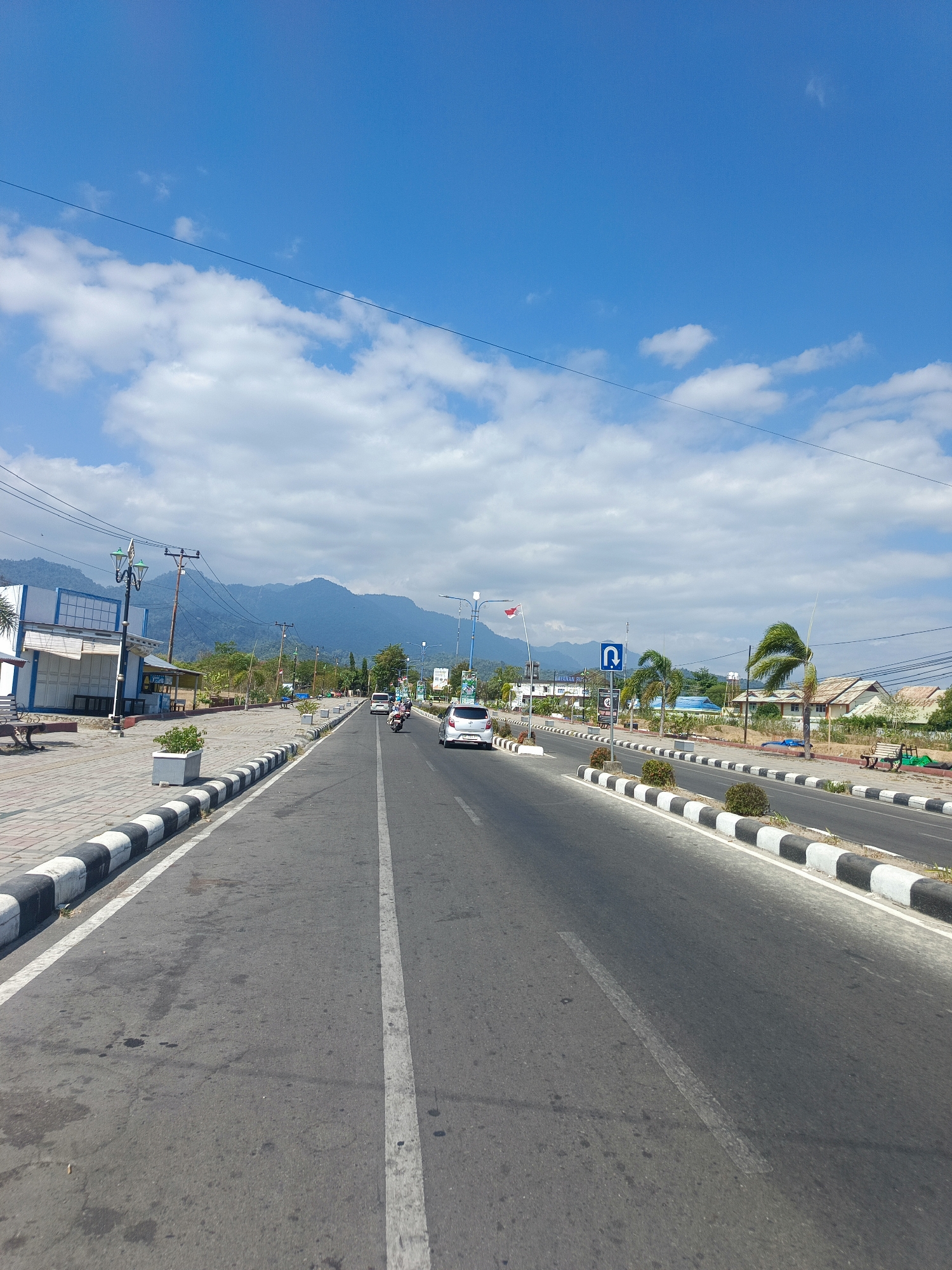 The image depicts a street scene in Suwawa, Indonesia, under a clear blue sky with scattered clouds. A paved road dominates the foreground, with white and black striped concrete barriers lining its edges. On the left side of the road, a sidewalk area features a building with white and blue facade, interspersed with street lamps and planters. Further down the road, several vehicles, including cars and motorcycles, are in motion. On the right side, houses and palm trees are visible, and a "U-turn" traffic sign is posted. In the background, a mountain range rises against the sky. The overall scene suggests a sunny day in a town with a blend of infrastructure and natural scenery.