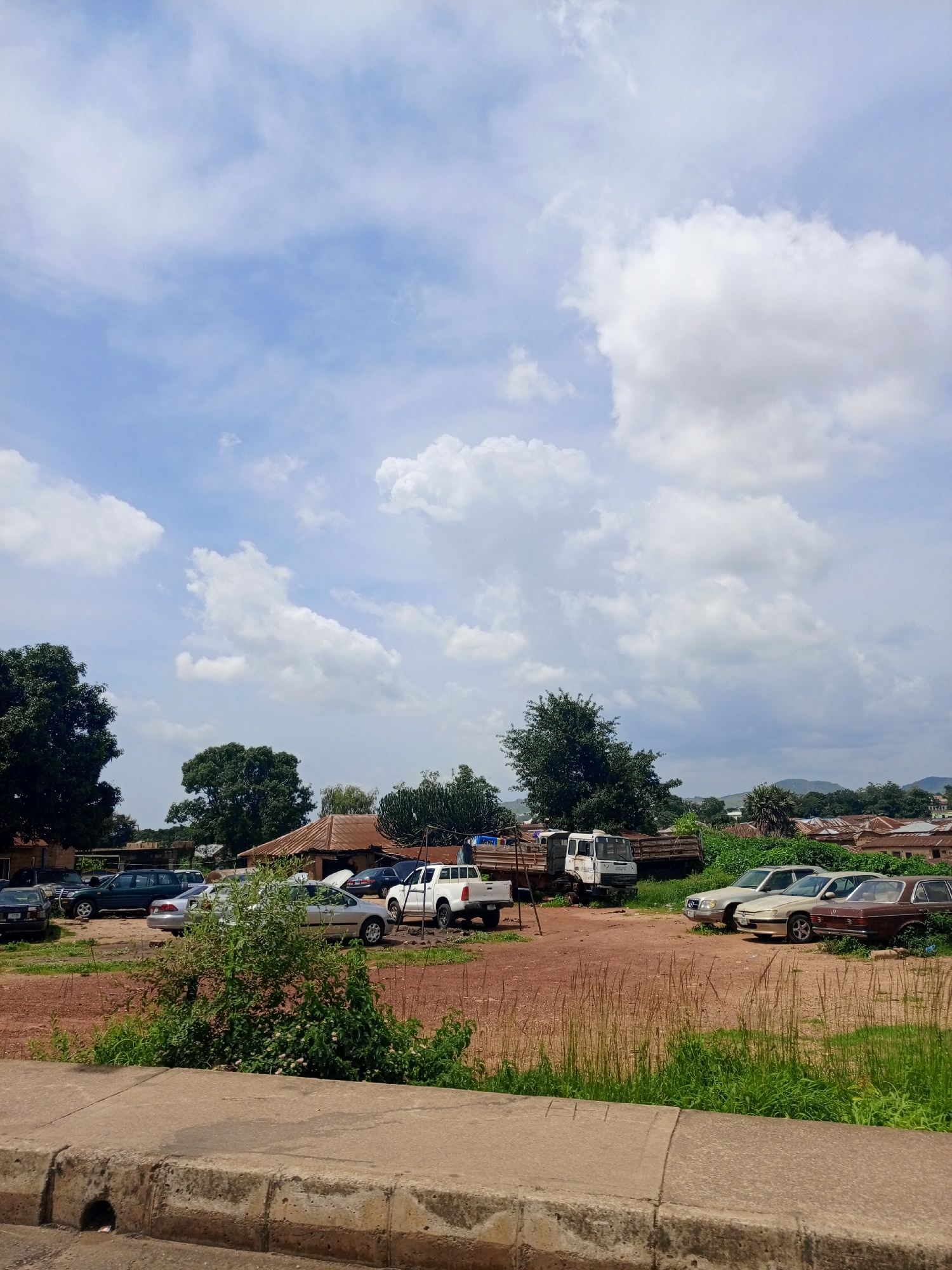 An outdoor, eye-level, full-shot image captures a parking lot with several vehicles in what appears to be a semi-urban or rural area, possibly in the city of New Stadium, Nigeria. The scene is dominated by a large, partly cloudy sky with bright white clouds scattered across a pale blue expanse.
In the foreground, a concrete curb runs horizontally across the bottom of the frame, suggesting the edge of a road or walkway. Beyond the curb, a patch of dry, reddish-brown dirt is visible, characteristic of unpaved ground. Tall, dry grass and green vegetation grow in clumps and patches throughout this area and spill onto the parking lot.
The parking lot itself contains a variety of vehicles. To the left, a dark-colored SUV is parked alongside other cars that are partially obscured. Further into the lot, a white pickup truck is parked prominently, facing towards the right. Behind it, two older model trucks are visible, one white and one brown, with their cargo beds loaded with what appear to be materials.
Several cars are parked in rows in the midground and right side of the frame. These include a silver sedan, a gold sedan, and a brown sedan, all appearing to be older models. They are positioned on the dirt lot, with some vegetation encroaching on them.
In the background, several buildings with corrugated metal roofs are clustered together, indicative of residential or commercial structures. Mature trees with dark green foliage are scattered throughout the scene, adding to the natural landscape. In the far distance, rolling hills are visible under the cloudy sky. The overall impression is of a somewhat utilitarian or perhaps neglected area, with a mix of vehicles and structures set against a natural backdrop. There are no people visible in the image.