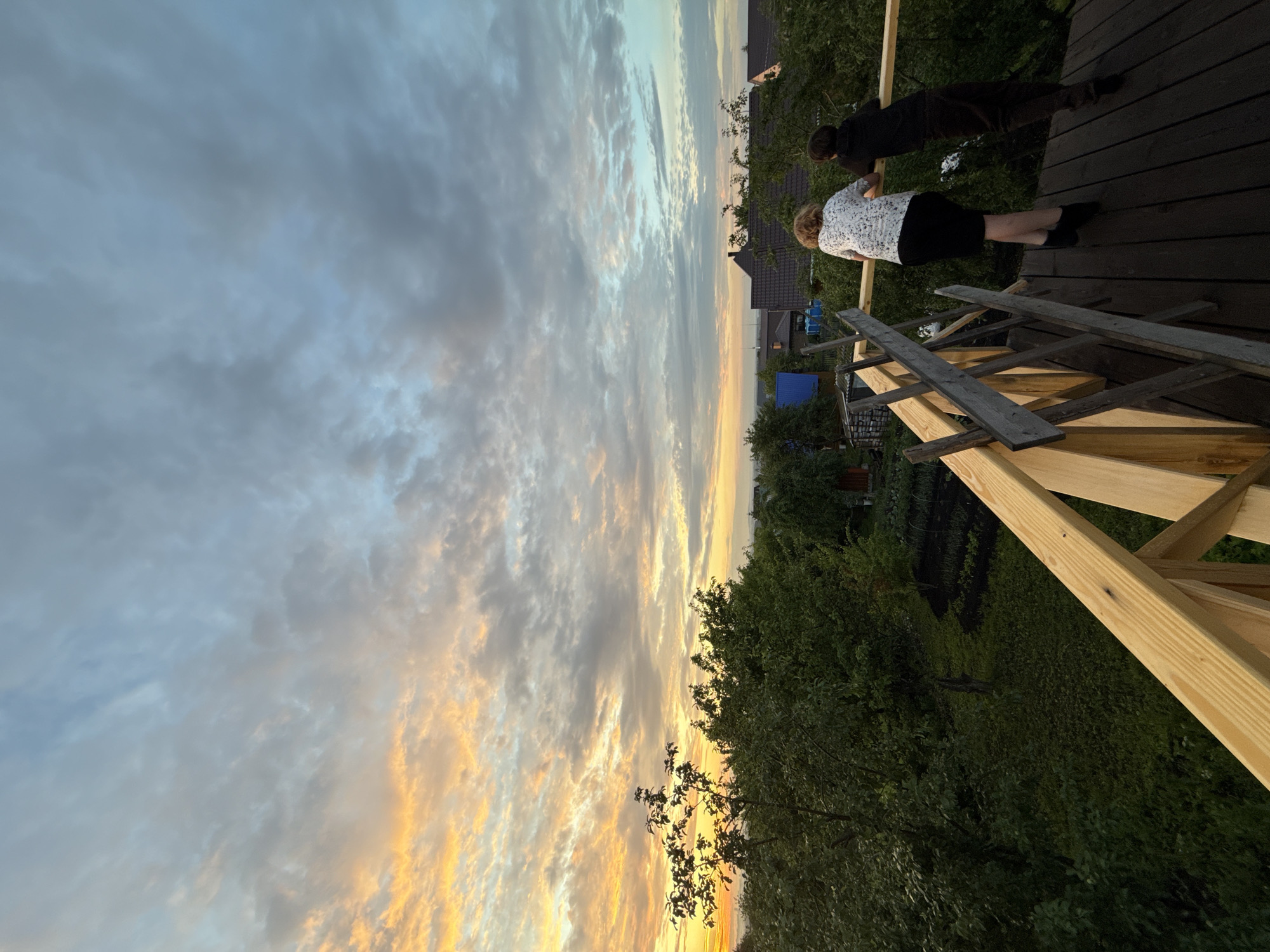 Two individuals, one with curly blonde hair and wearing a white patterned top and black skirt, and another wearing a dark hooded garment, stand on a wooden platform. They are looking out towards a landscape at dusk. The sky above is filled with clouds, with streaks of orange and yellow light from the setting sun visible on the left side, transitioning to cooler blue and gray tones on the right. Below the platform is a green, leafy landscape with trees and what appear to be garden beds. Several rooftops of houses are visible in the mid-ground, suggesting a rural or suburban setting. The platform itself appears to be under construction, with unfinished wooden railings.