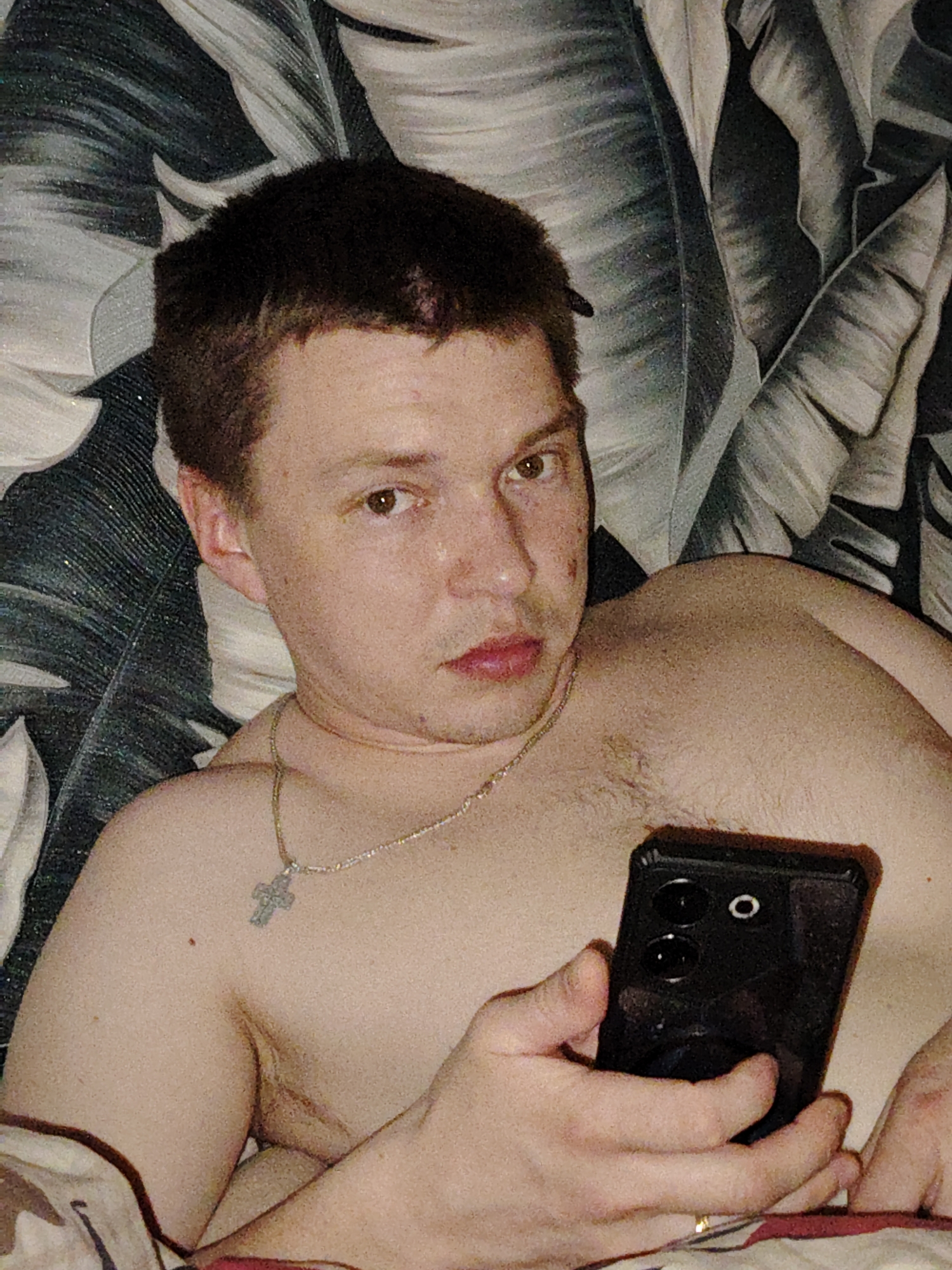 A young adult male is depicted in an indoor, close-up shot, reportedly in Марковский, Russia, though the precise geographical location is not visually verifiable from the image. The male has short, dark hair and brown eyes, gazing directly towards the camera with a neutral expression. He appears shirtless, exposing his upper chest and shoulders, which bear light body hair. Around his neck, a thin, silver-colored chain with a small crucifix pendant is visible. His left hand, wearing a thin gold-colored ring on the ring finger, is partially visible. His right hand holds a black smartphone vertically against his chest; the phone features a distinct camera module with two prominent circular lenses and a smaller circular flash or sensor. The background consists of a patterned fabric, likely bedding or upholstery, displaying large, stylized leaf motifs in shades of dark grey, light grey, and white.
